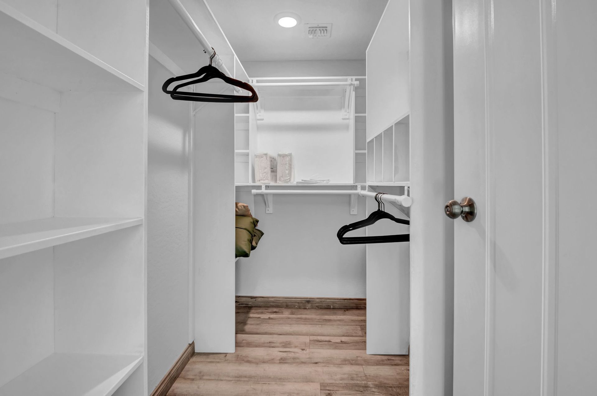 White walk-in closet with shelves, hanging rods, and wood-look flooring. Door open, two hangers visible.