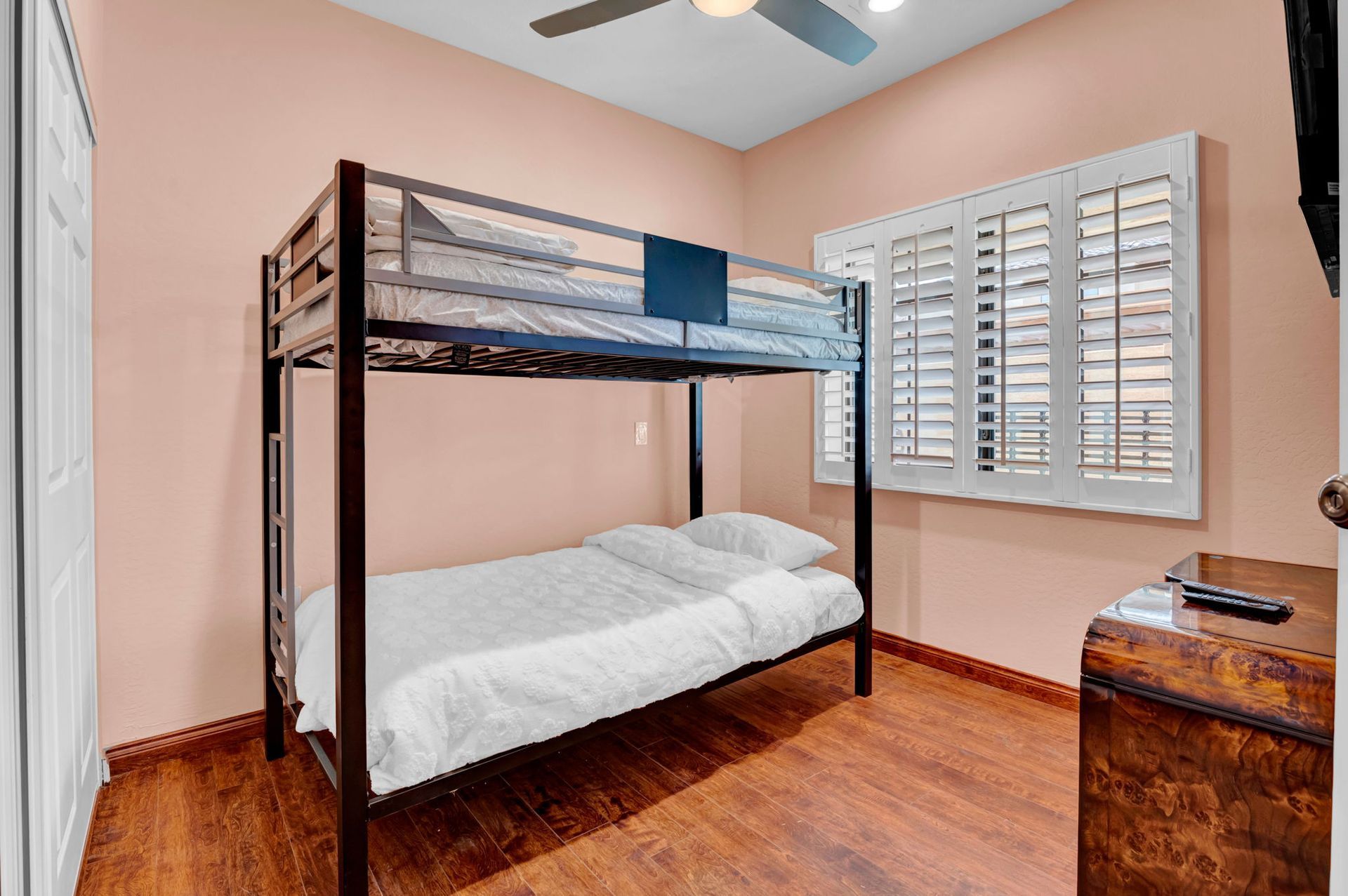 Bunk beds in a room with wood flooring, pink walls, and a window with shutters. There's also a trunk.