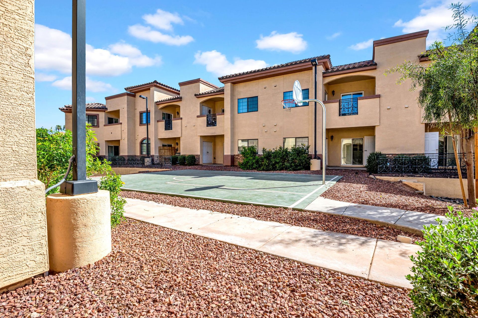 Apartment building exterior with a basketball court. Beige stucco, blue sky, and red landscaping.