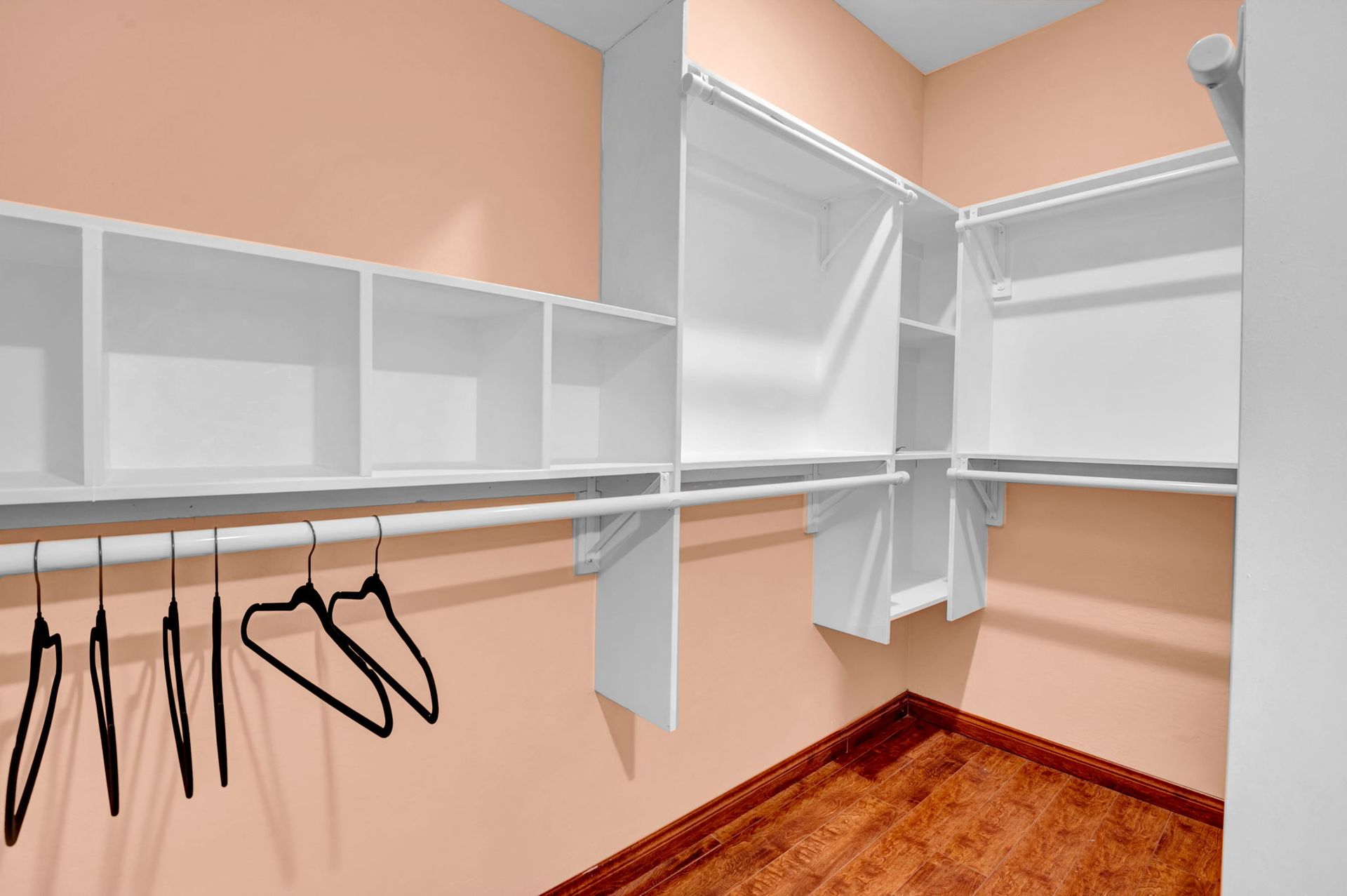 Empty walk-in closet with white shelving and rods, peach walls, and wood floor. Black hangers hang from a rod.