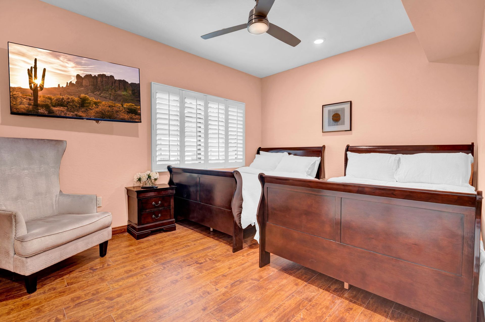 Bedroom with two beds, armchair, TV, window with shutters, wood floors, peach walls.