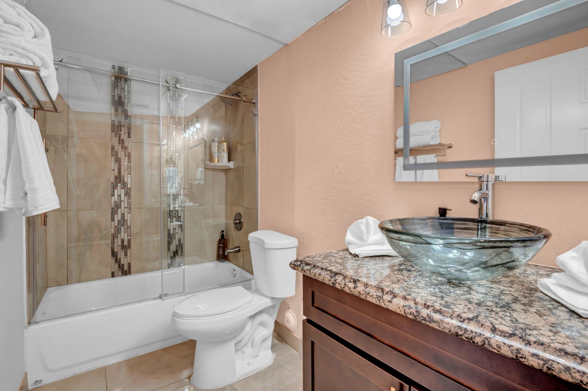 Bathroom with a glass bowl sink on a granite countertop, toilet, bathtub with glass shower doors, and towels.