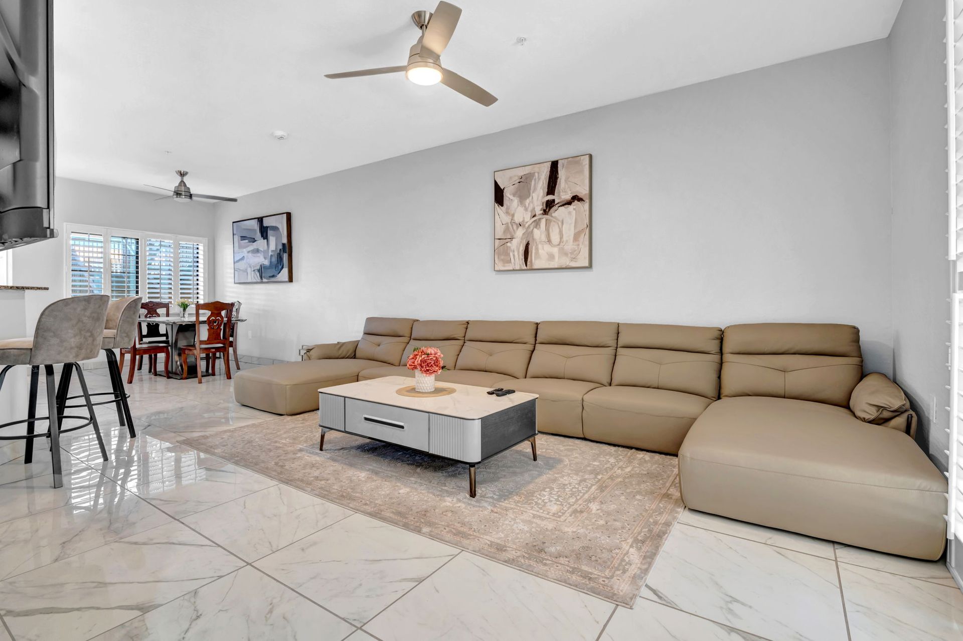 Living room with beige sectional sofa, coffee table, rug, and artwork. Dining area in the background.