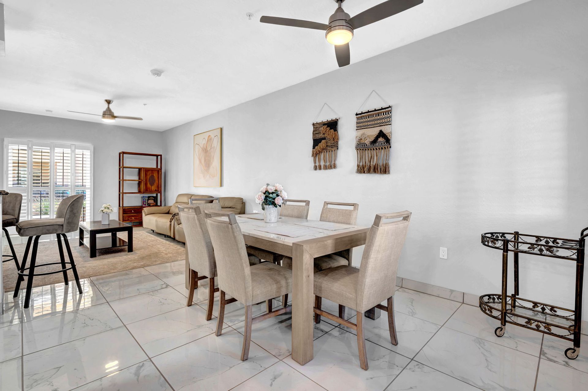 Dining area with table, chairs, bar stools, and artwork on the wall. Open to living room with couch and shelving.