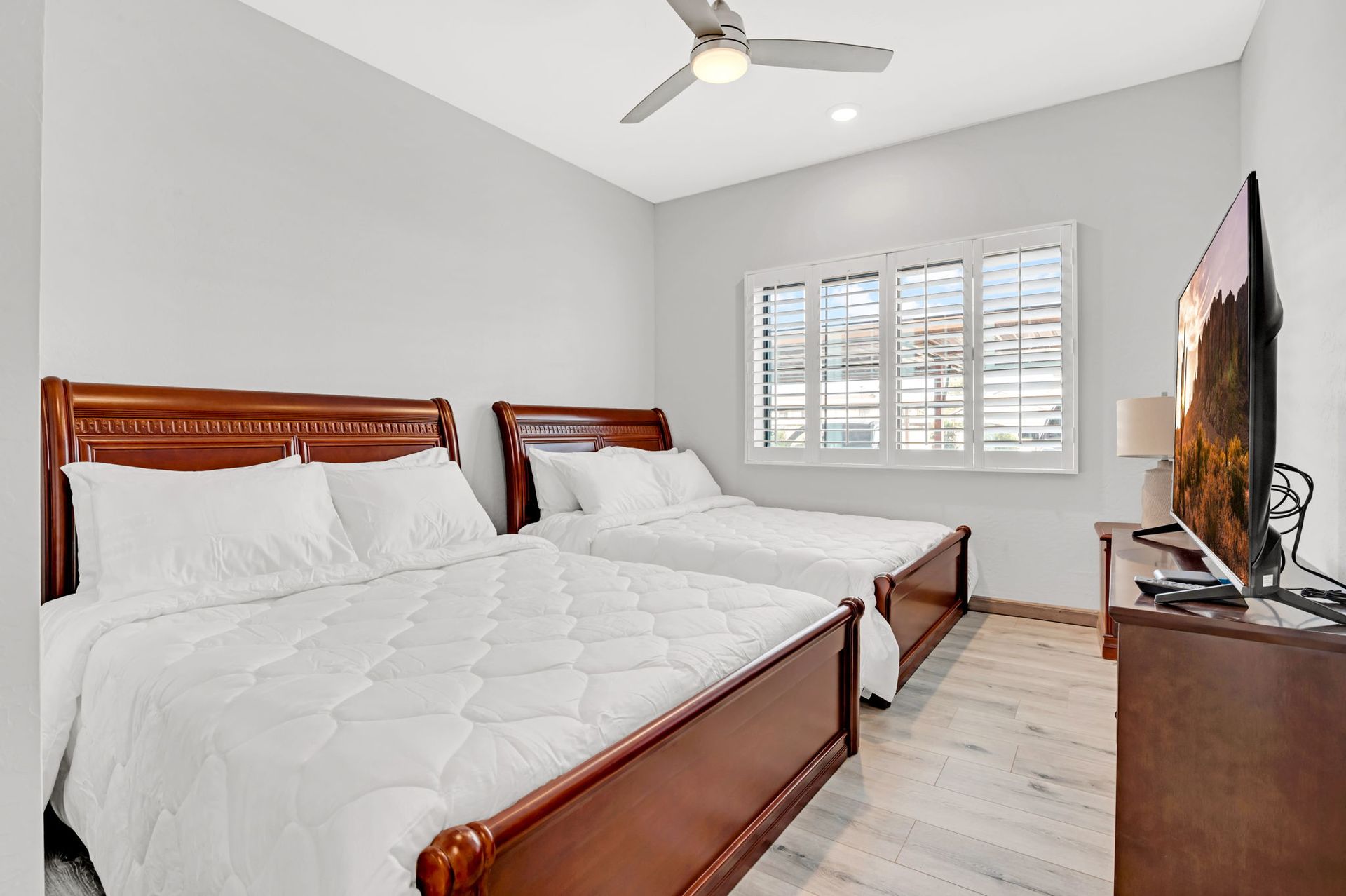 Bedroom with two beds, wooden frames, white bedding, shuttered window, TV, ceiling fan.