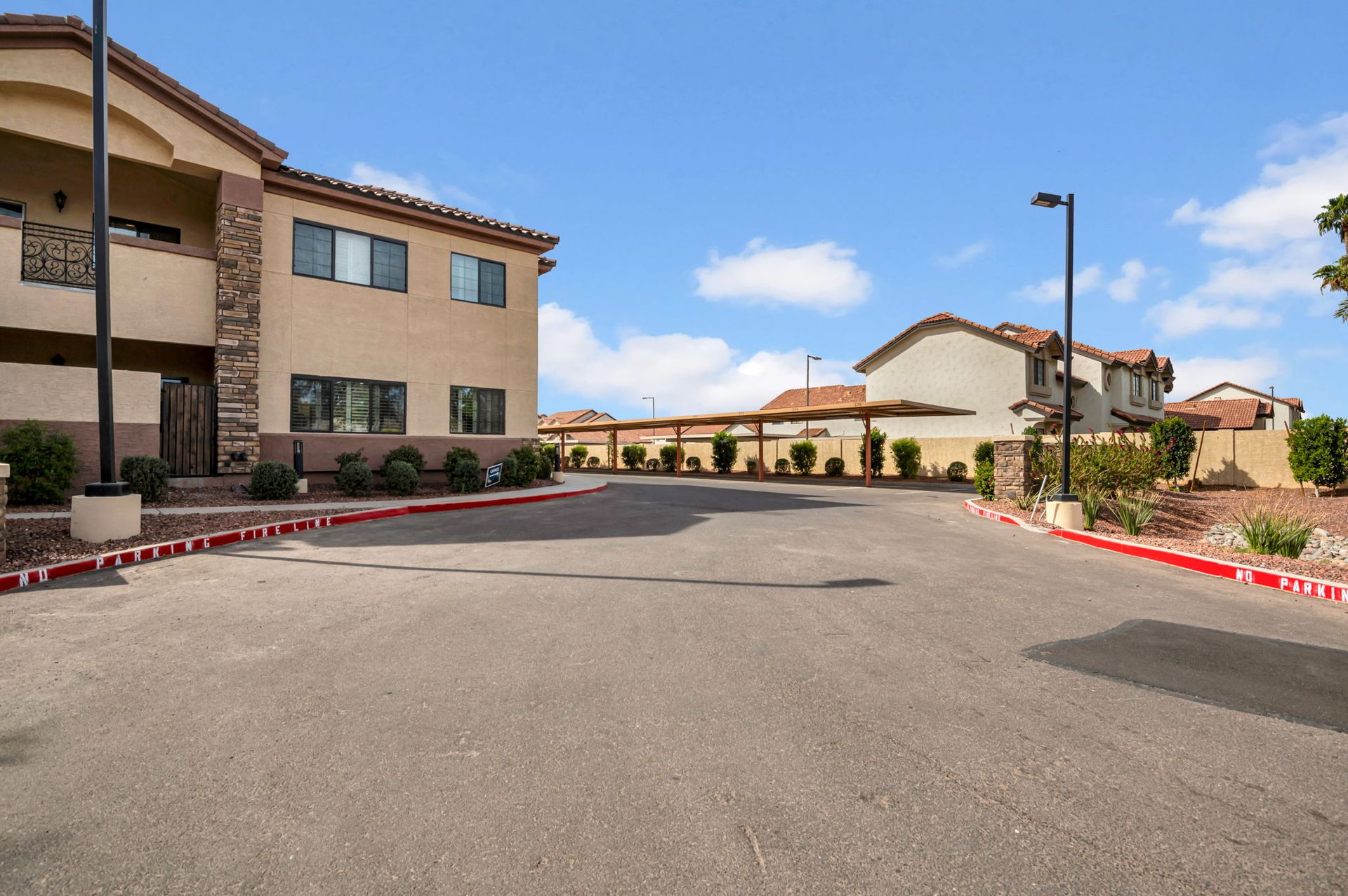Asphalt driveway leading past tan multi-story buildings and covered parking under a blue sky.