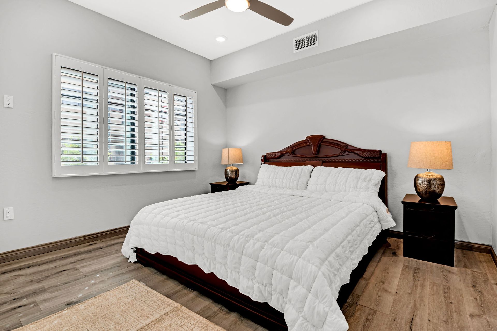 Bedroom with bed, nightstands, lamps, window with shutters, and wood flooring.