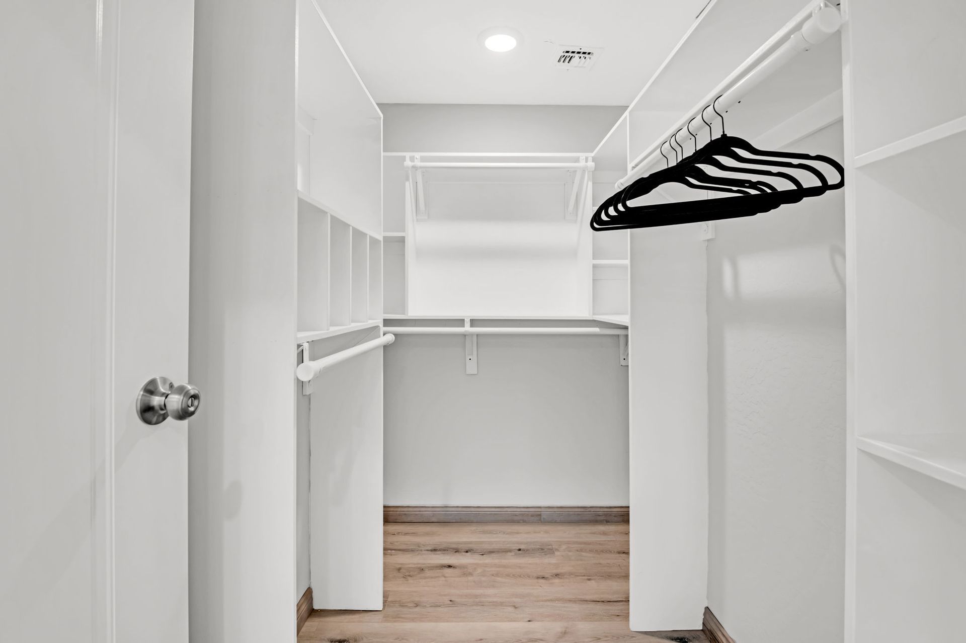 Empty, white walk-in closet with shelves, hanging rods, and a door, bright lighting, and wood flooring.