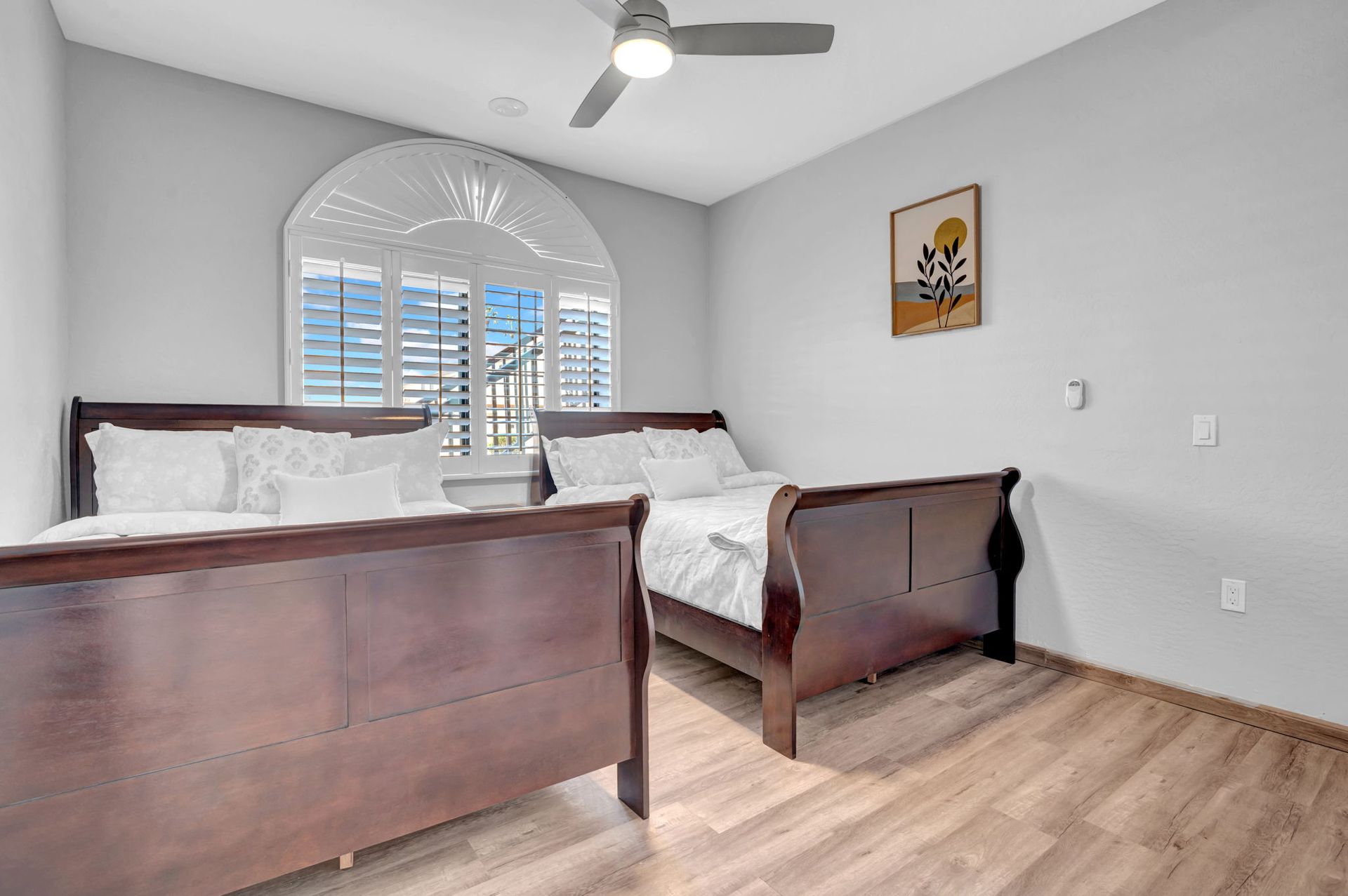 Bedroom with two brown sleigh beds, light wood floors, and arched window with shutters.