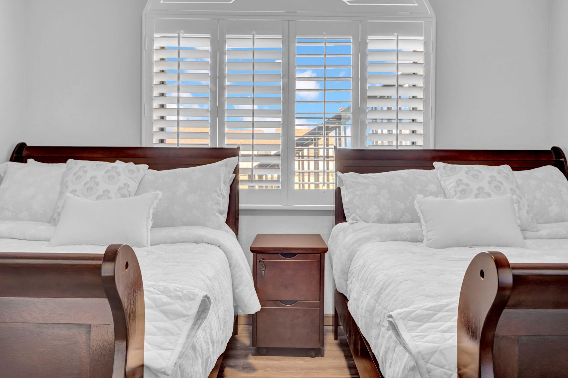 Two twin beds with white bedding, flanked by a dark wood nightstand, in a room with a shuttered window.