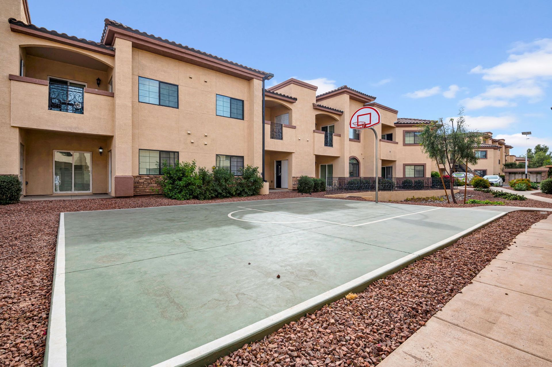 Basketball court in front of beige apartment building with bushes, brown stones, and blue sky.