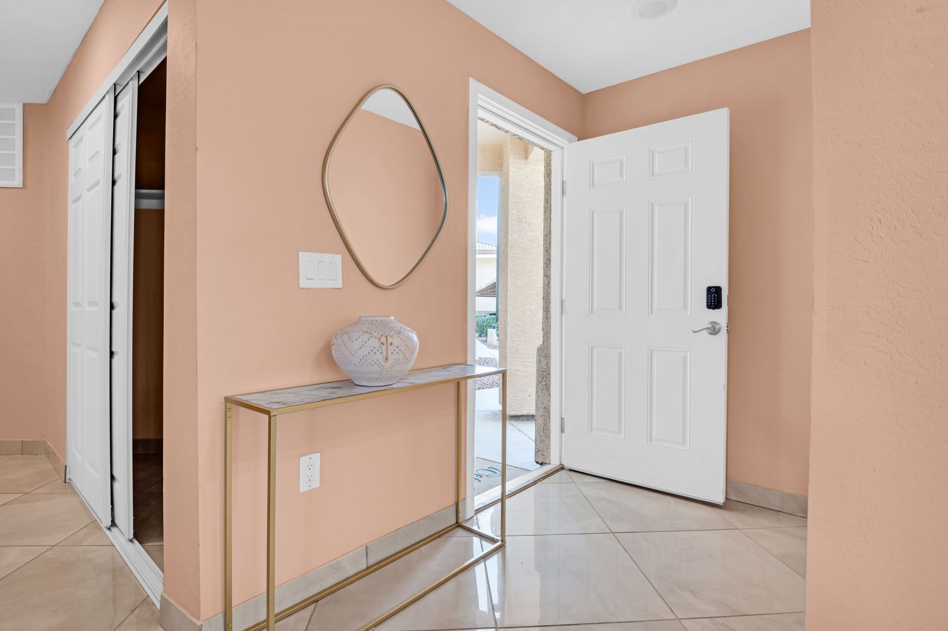 Peach-painted entry with open white door, closet, gold console table, mirror, and decorative vase.