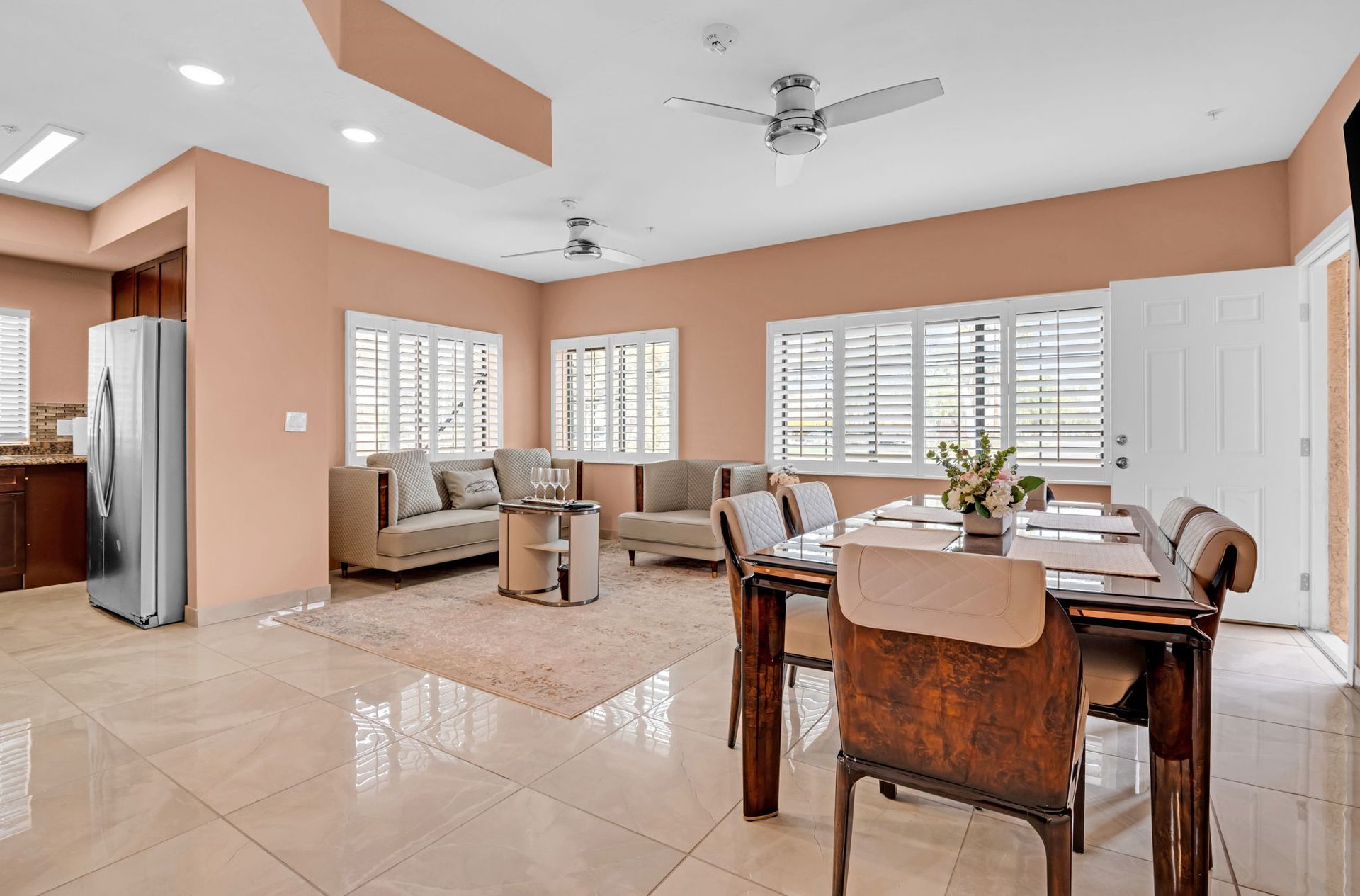 Open-concept living space with dining area, sofas, and kitchen. Light peach walls, white shutters, tile floor.