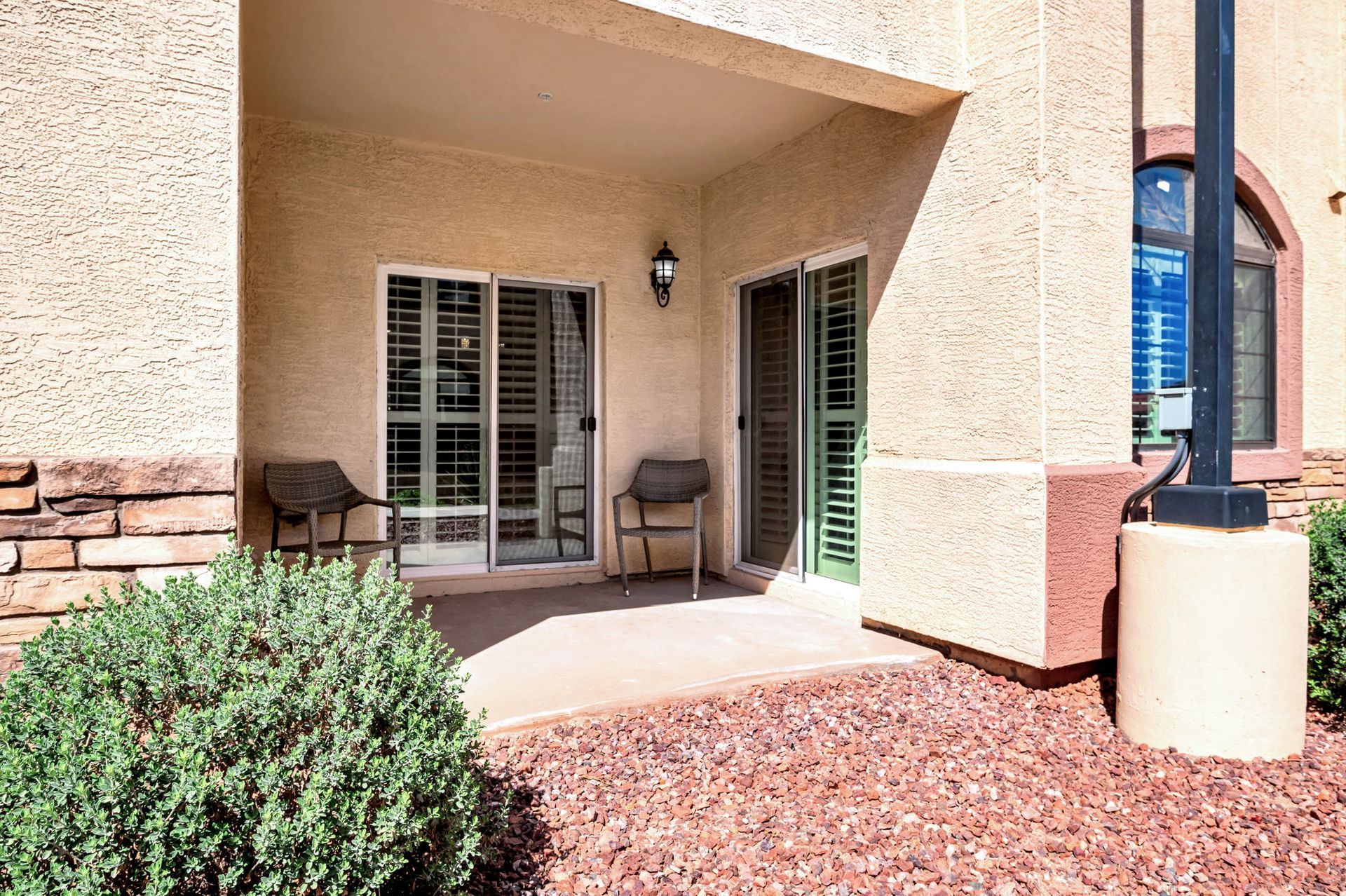 Patio with sliding doors, chairs, and shrubbery; brown gravel ground and beige stucco building.