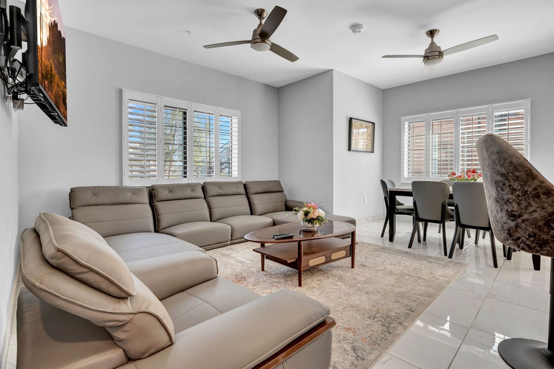 Living room with grey sectional sofa, dining table, shutters, and ceiling fans.