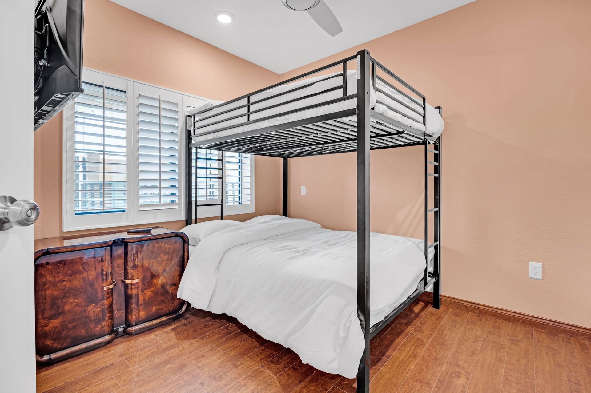 Bedroom with a black bunk bed, window with white shutters, and wood-like flooring. Peach walls and brown cabinet.