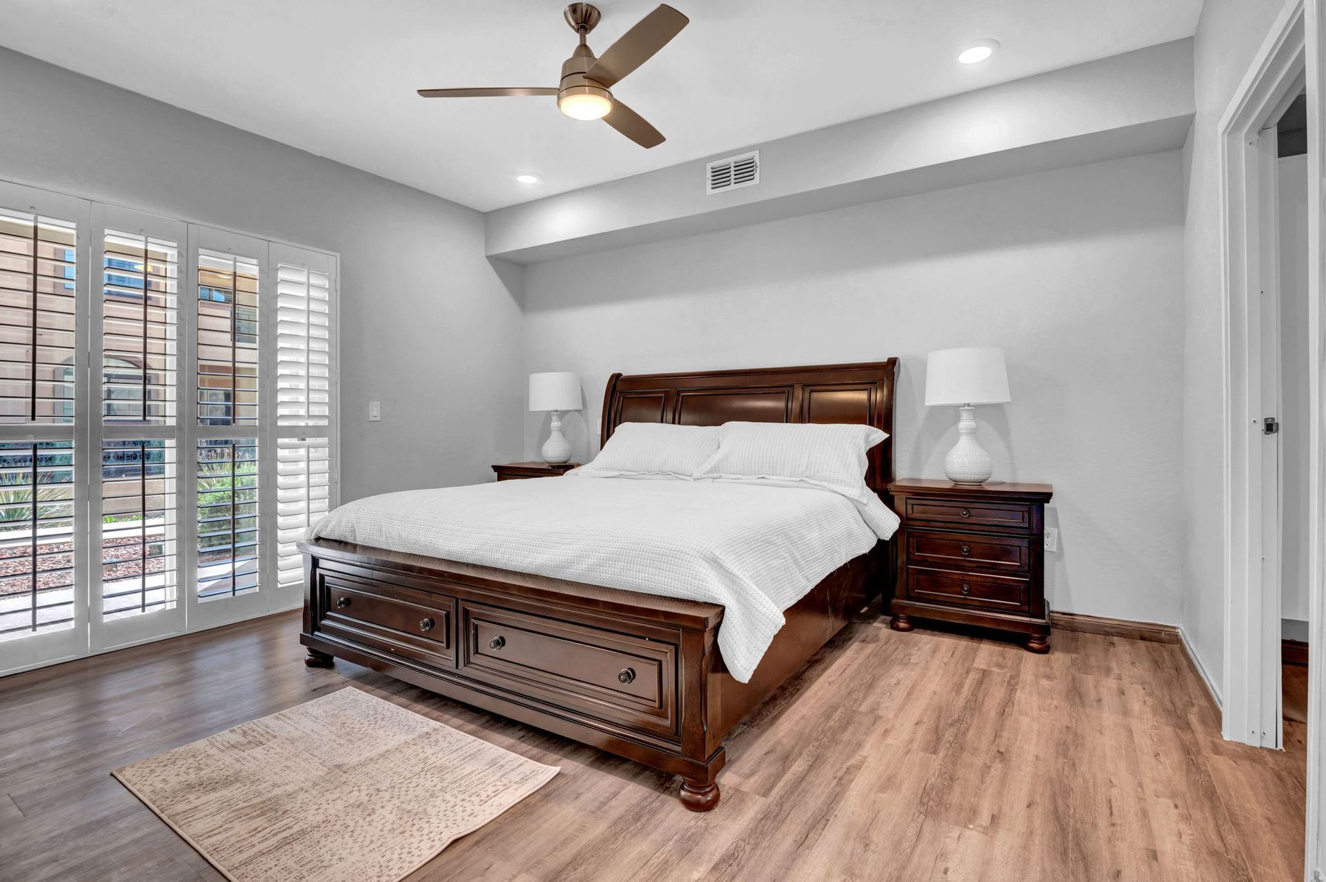 Bedroom with wooden bed, nightstands, and window with shutters. Pale gray walls, light brown floors.