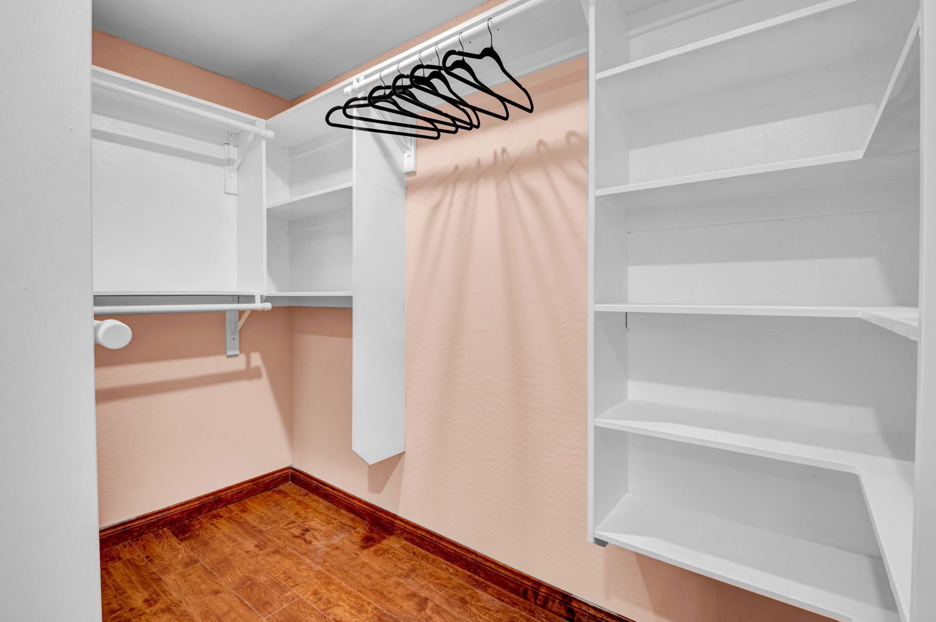 Empty white closet with built-in shelves and hanging rod on a wood floor, with pink walls.