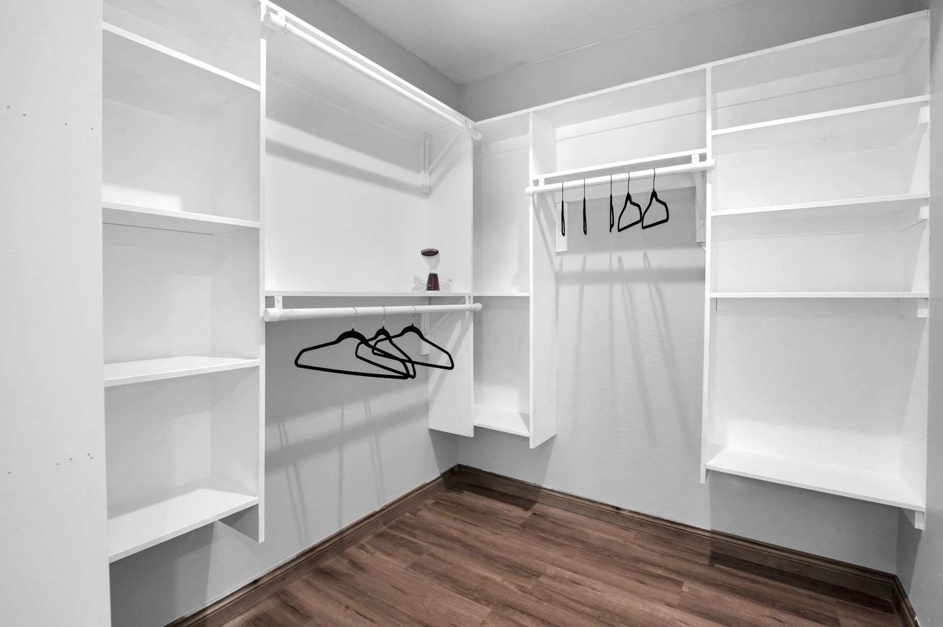 Empty walk-in closet with white shelves, hanging rods, and dark wood-look floor.
