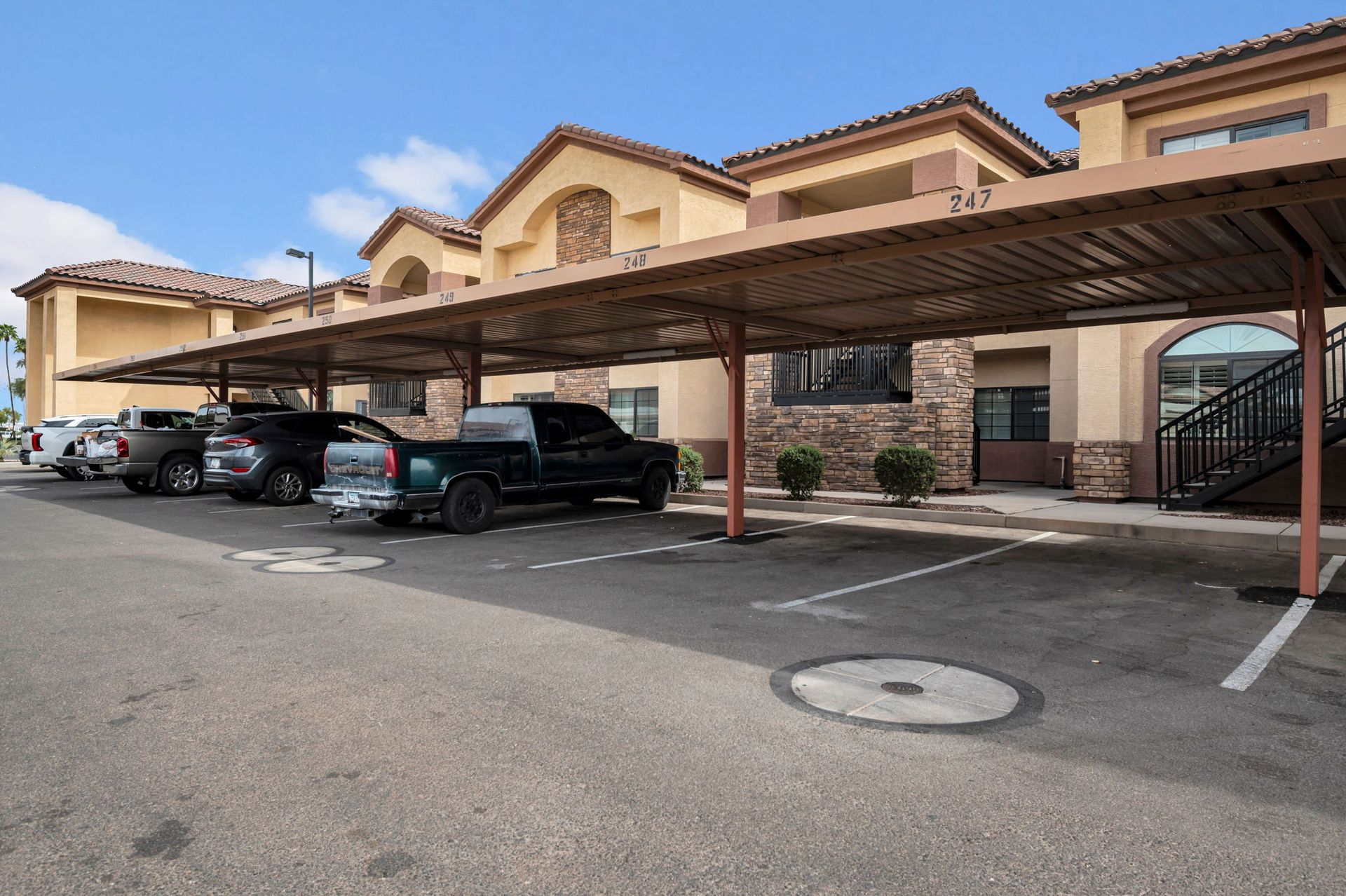 Covered parking lot with parked cars in front of a tan apartment building under a blue sky.