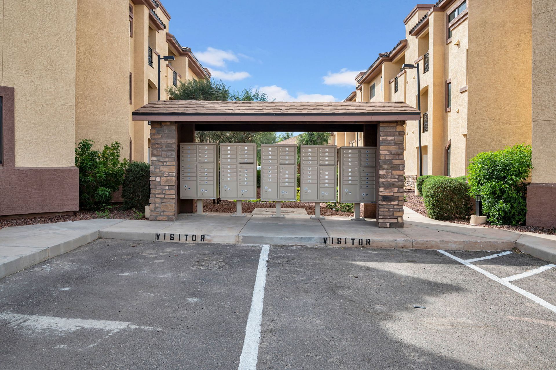 Mailboxes under a covered structure in front of apartment buildings. Asphalt parking lot.
