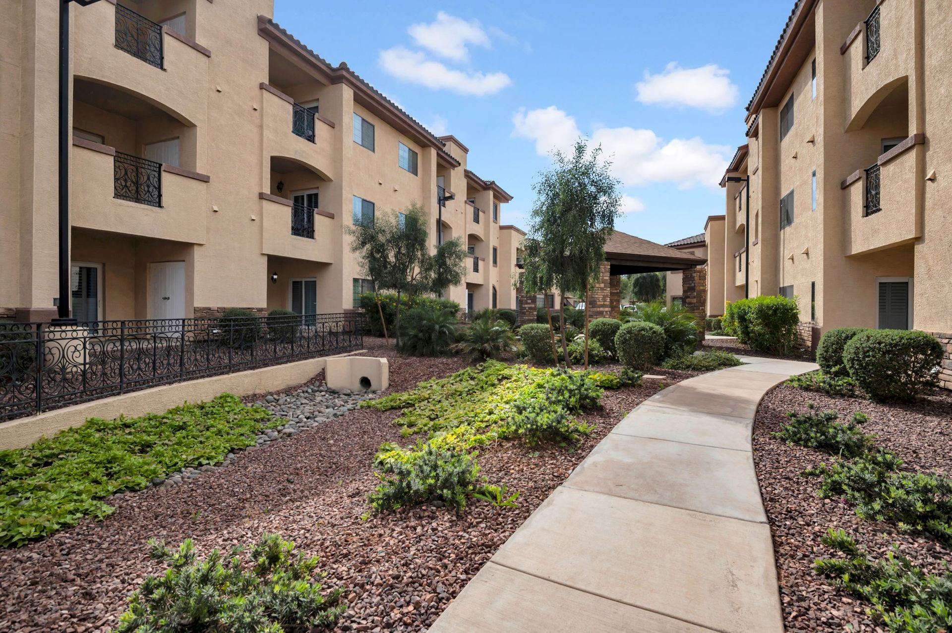 Apartment complex with a stone walkway, landscaping, and a blue sky. Buildings are tan with black railings.