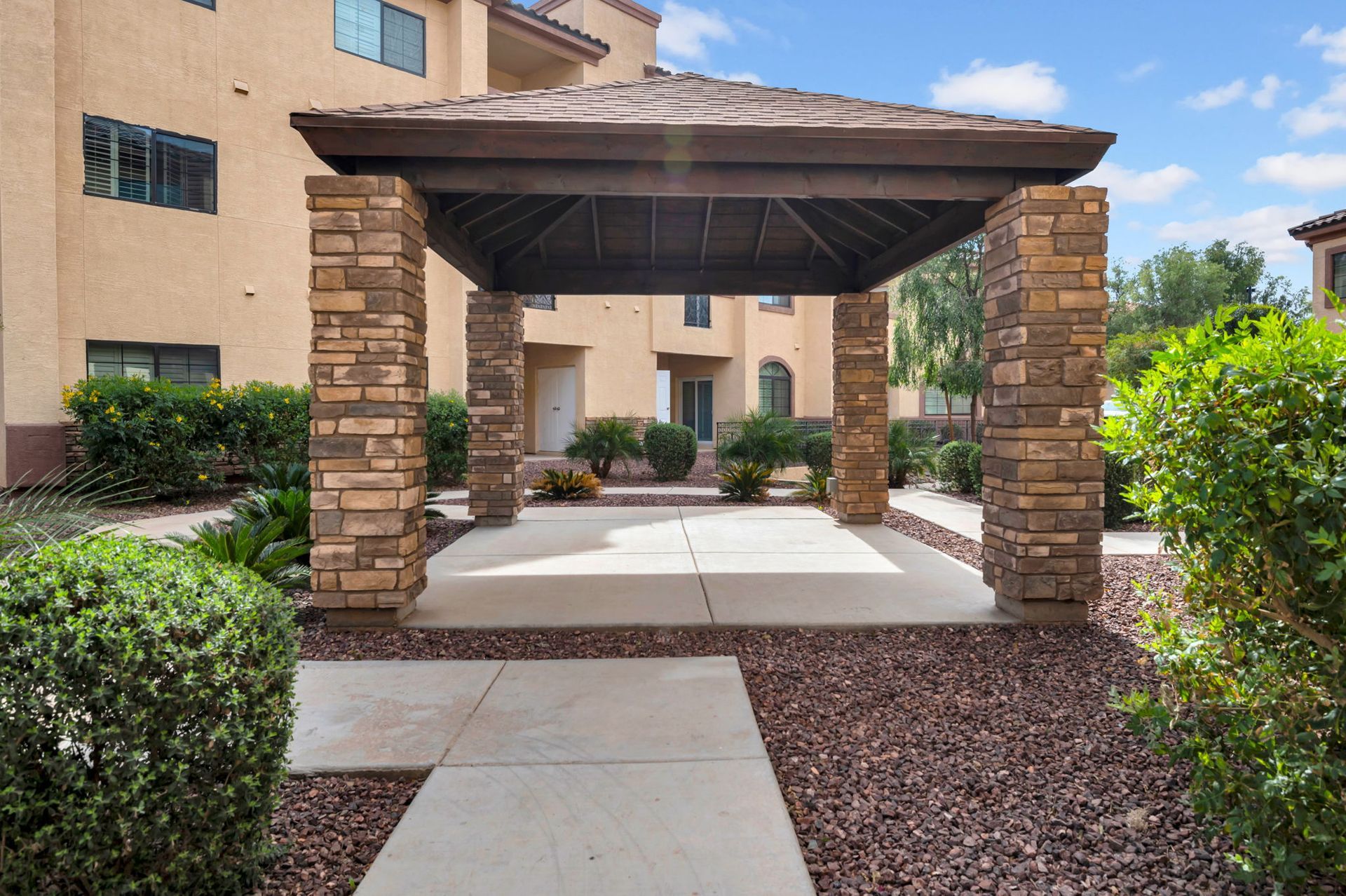 Covered patio with stone columns and a concrete floor, in a landscaped area near a building.