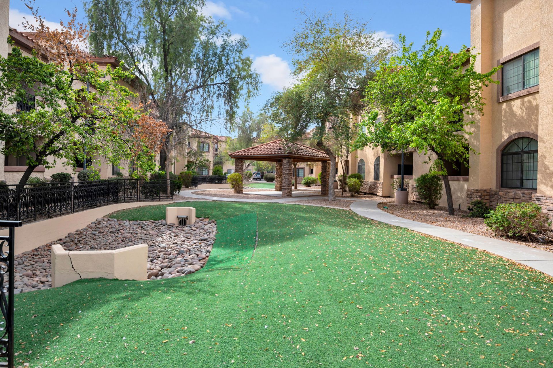 Courtyard with artificial turf, stone gazebo, trees, and tan buildings.