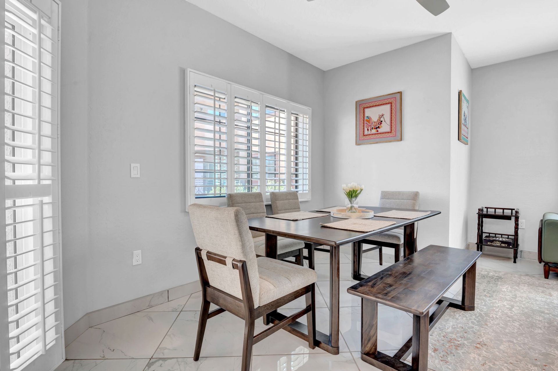 Dining room with a table, chairs, bench, window with shutters, and artwork.