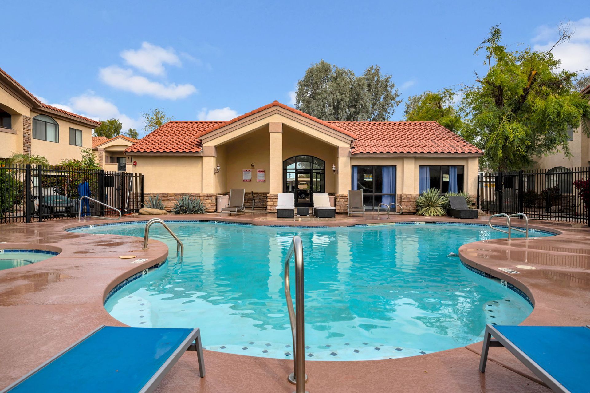 Pool with lounge chairs in front of a tan building with a red-tiled roof, under a blue sky.