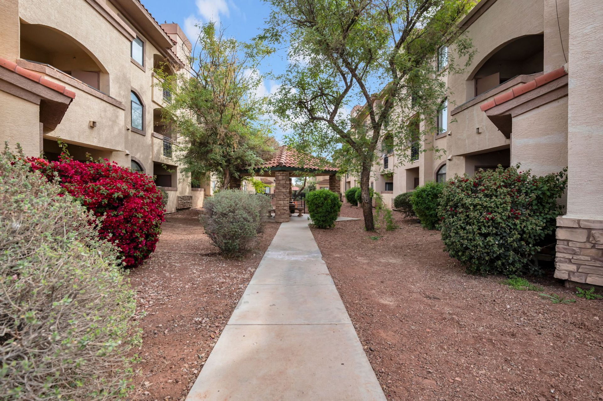 Pathway between apartment buildings, with red and green bushes, and trees under a cloudy blue sky.