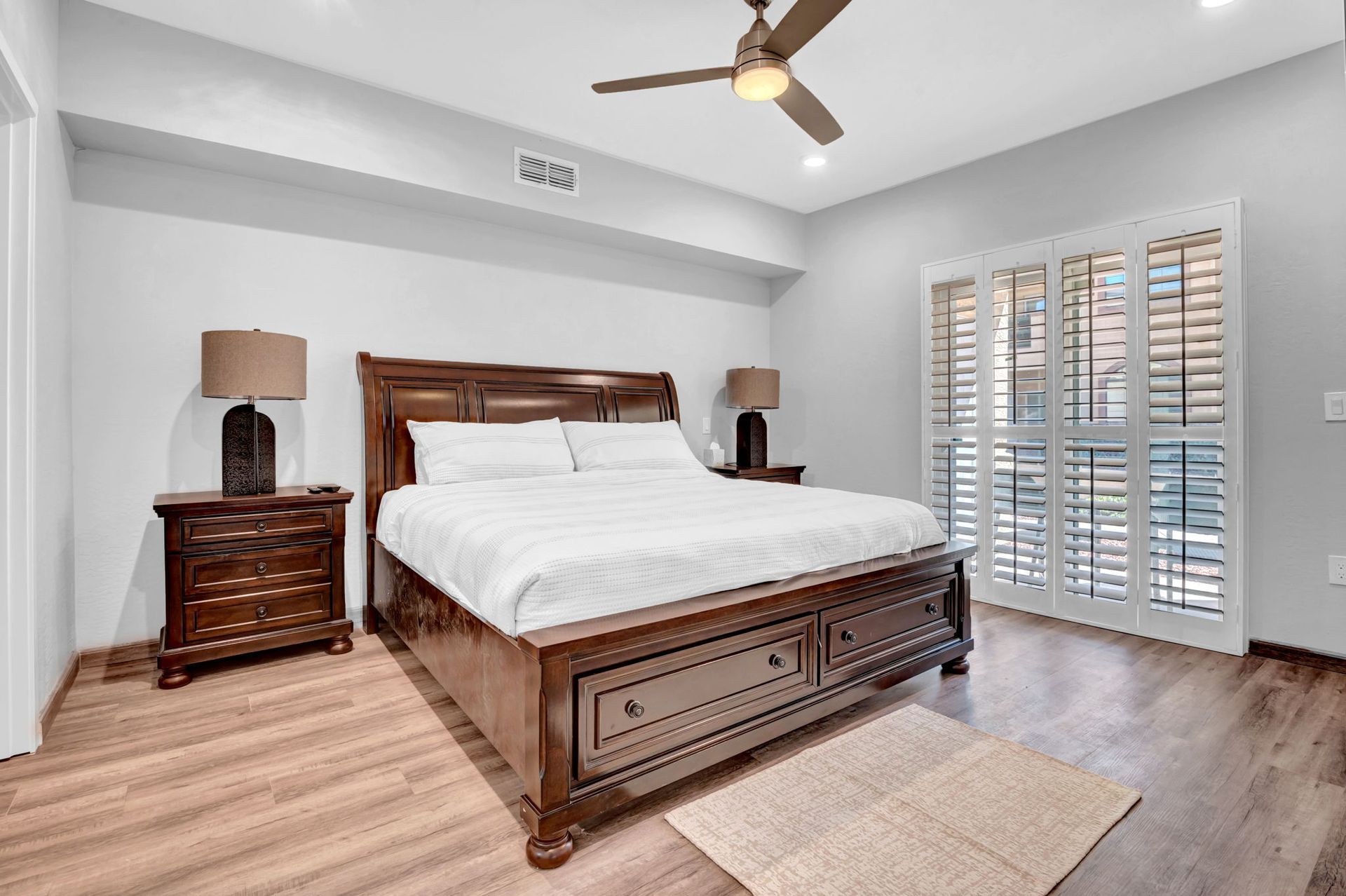Bedroom with a large bed, nightstands, lamps, window shutters, ceiling fan, and hardwood floor.
