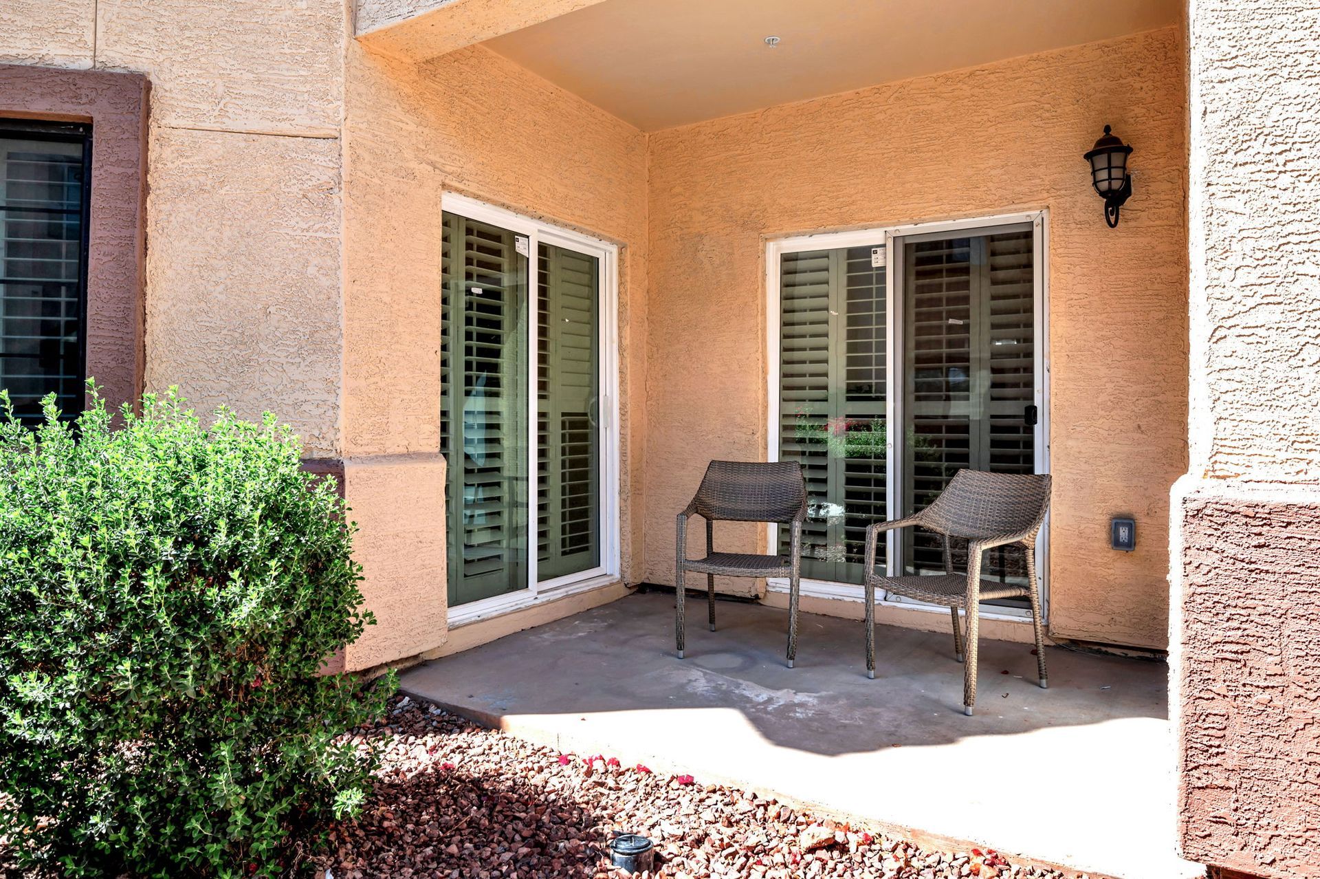 Patio with two chairs, two windows with shutters, and a bush. The walls are stucco.