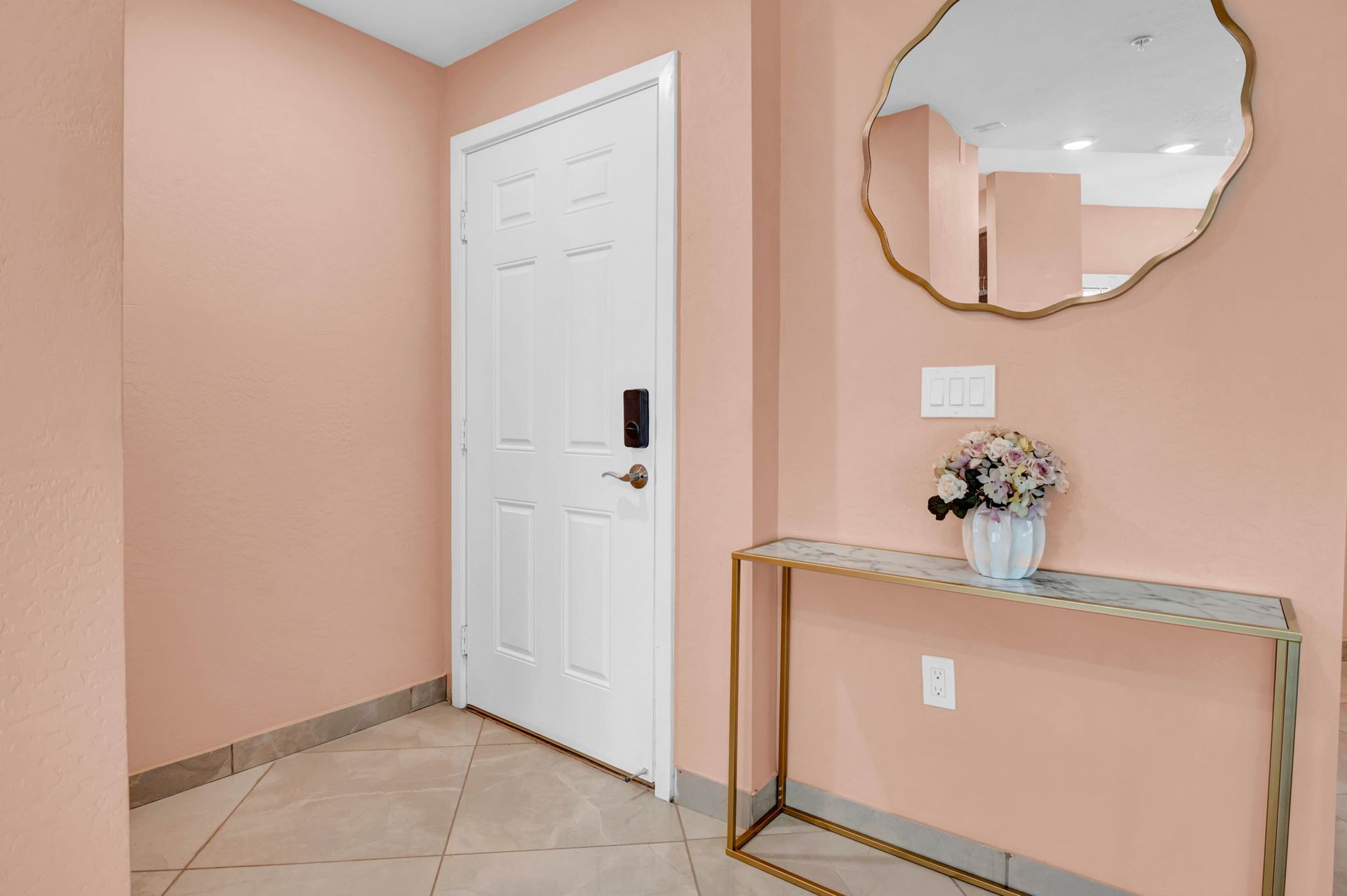 Pink entryway with white door, gold-legged table, and a decorative mirror and vase of flowers.