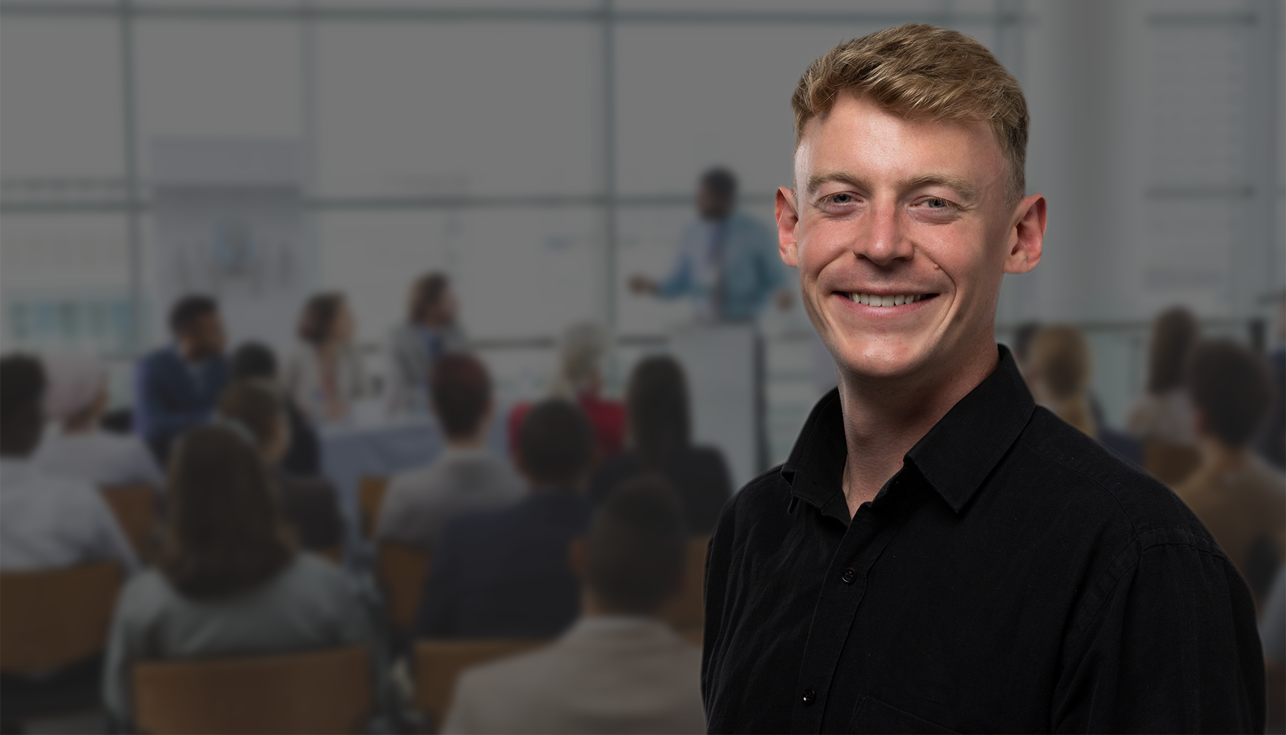 Man smiles, black shirt, in front of blurred audience and speaker at a presentation.