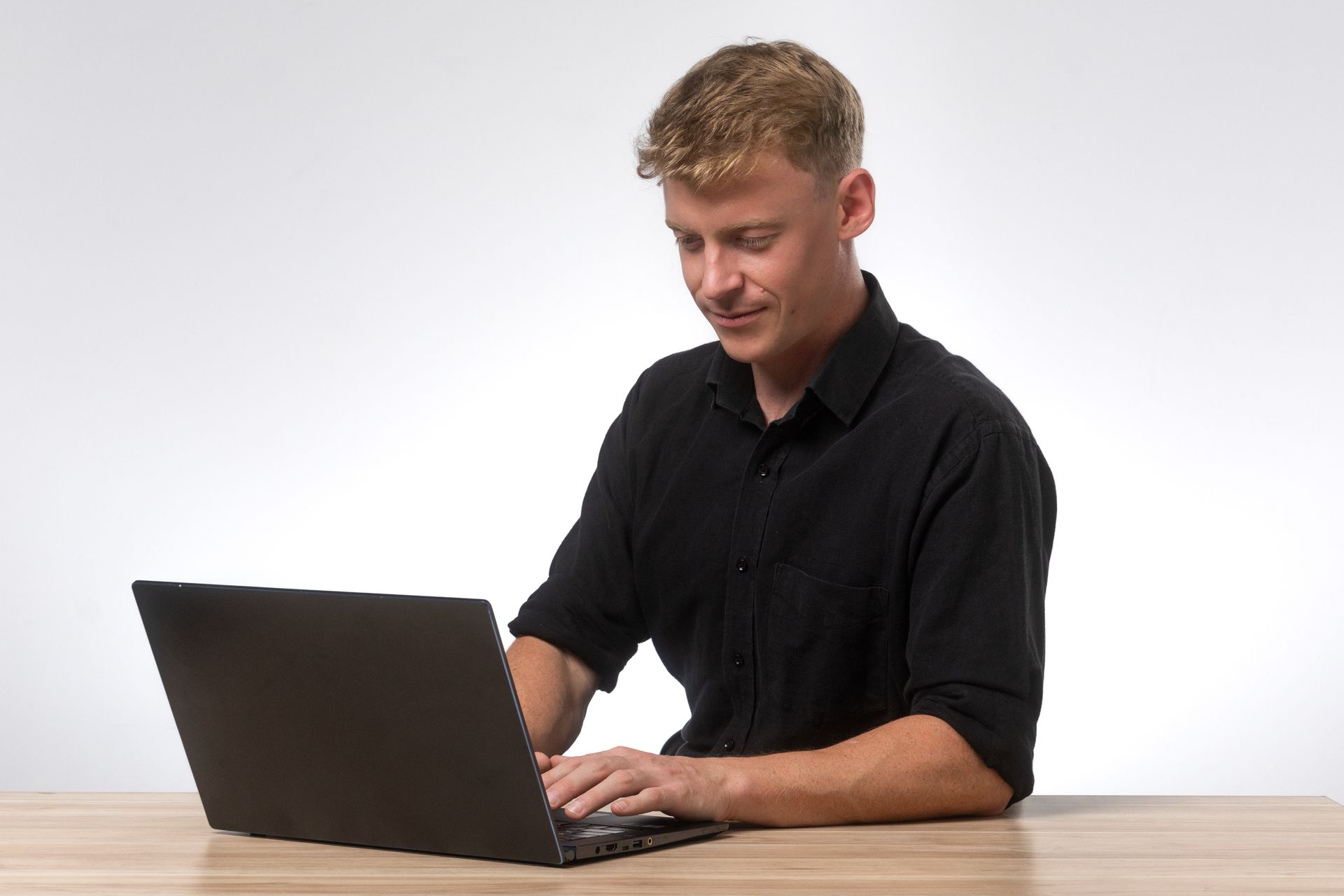 Man typing on laptop, seated at a wooden desk, wearing a black shirt, smiling.