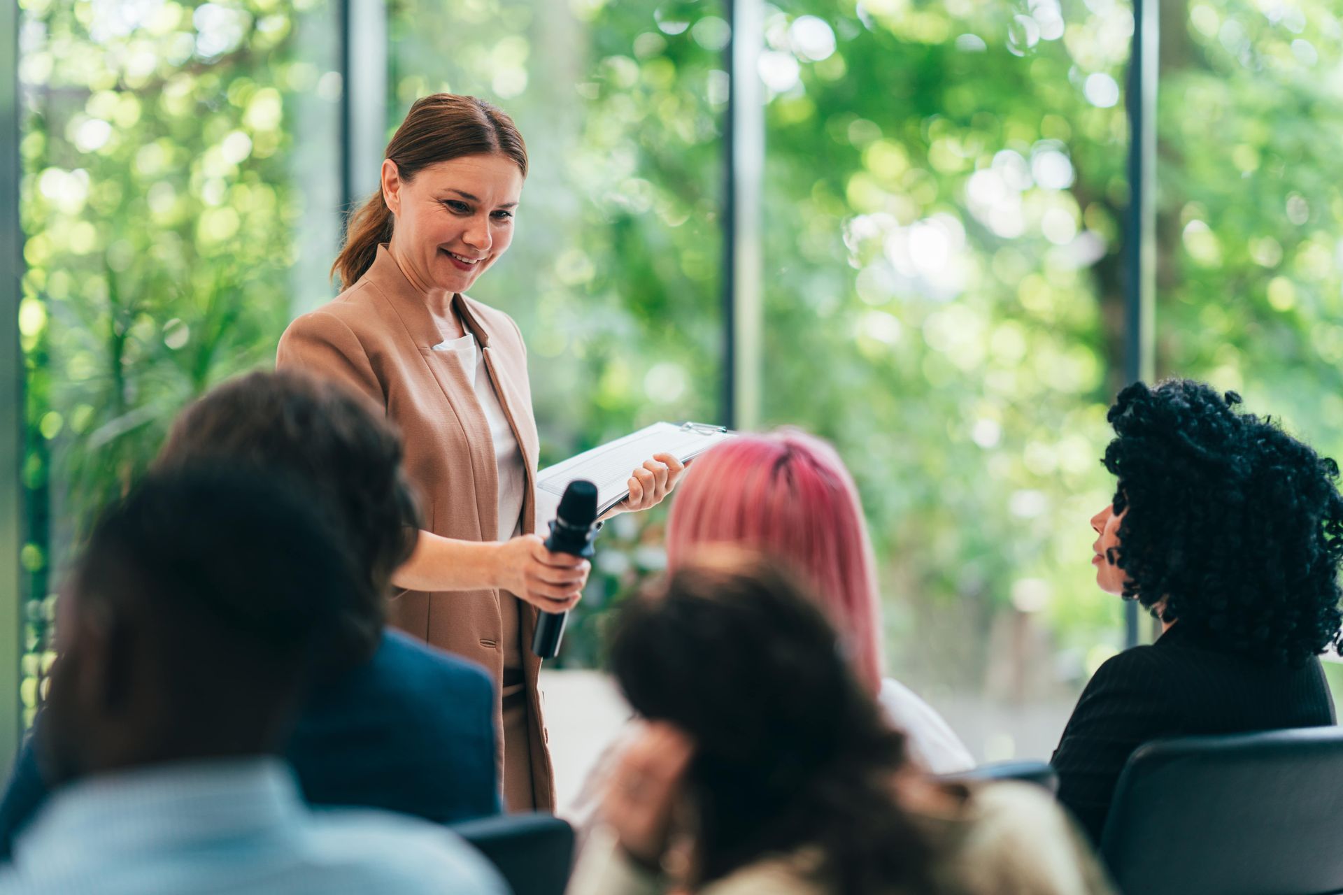 Woman speaking into a microphone, holding papers, facing an audience. Conference room setting with trees visible.