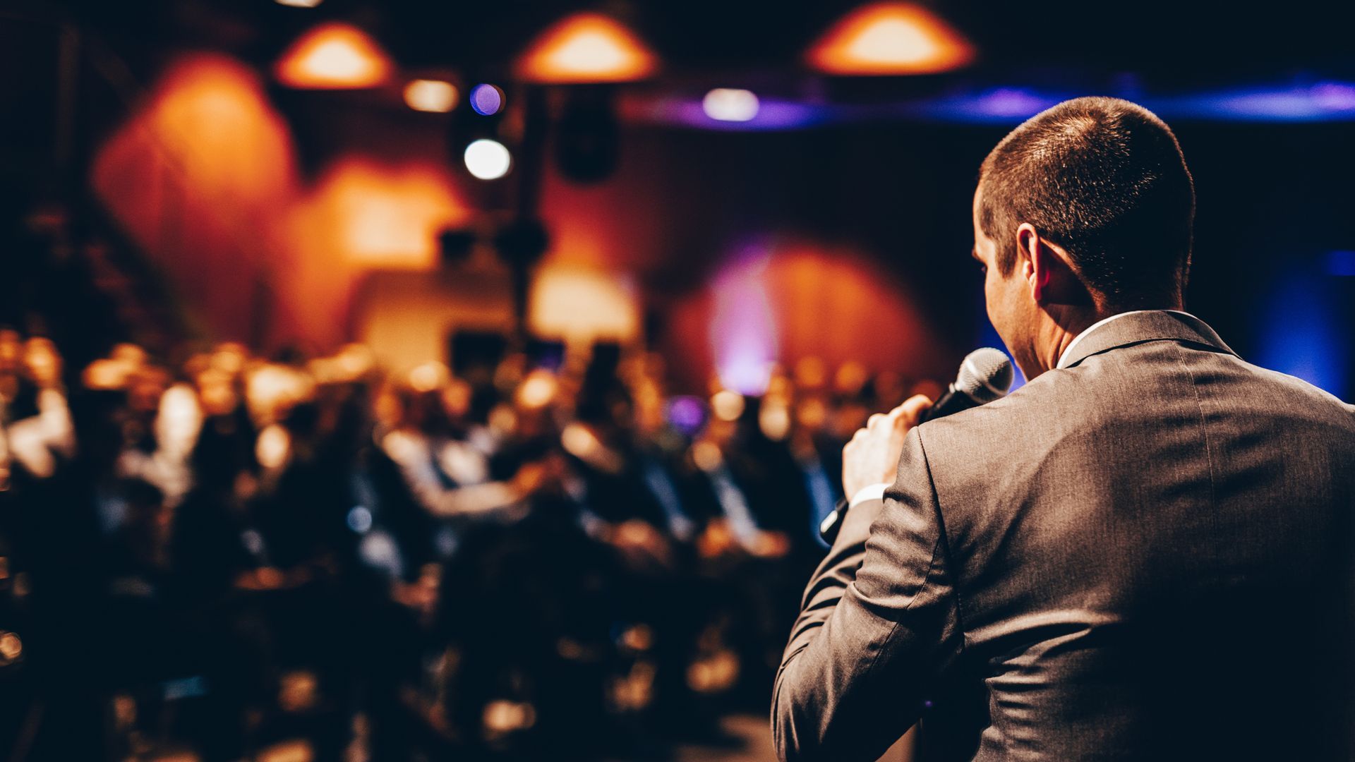 Man in suit speaking into microphone on stage, facing a blurry audience.