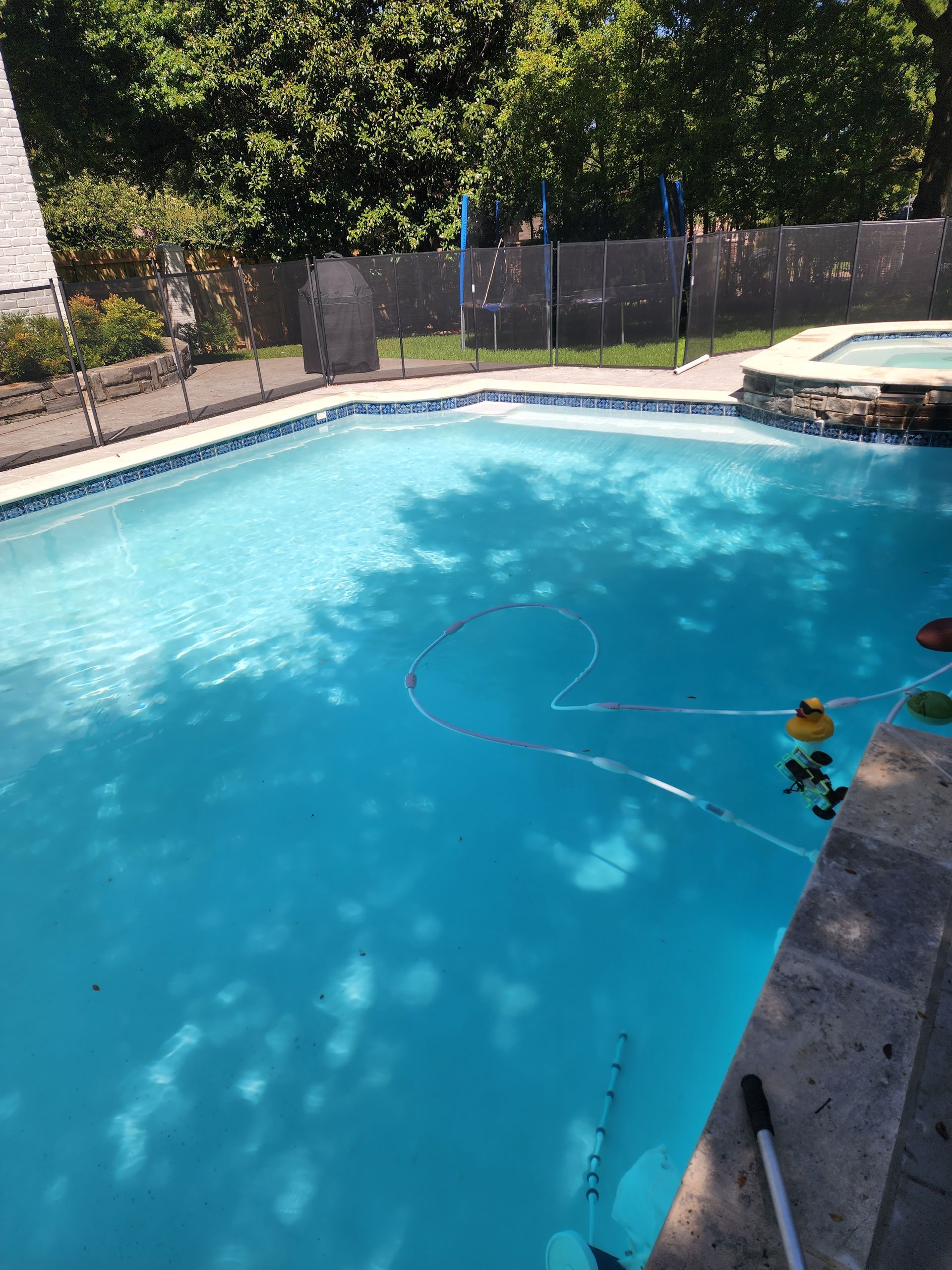 Blue swimming pool with safety fence, surrounded by a stone deck and trees, sunny day.