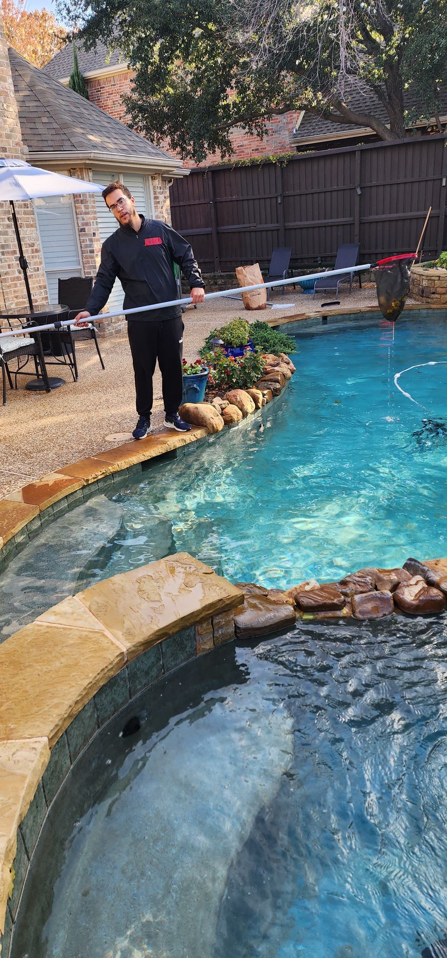 A man cleaning a blue pool with a long-handled net, standing on the edge.
