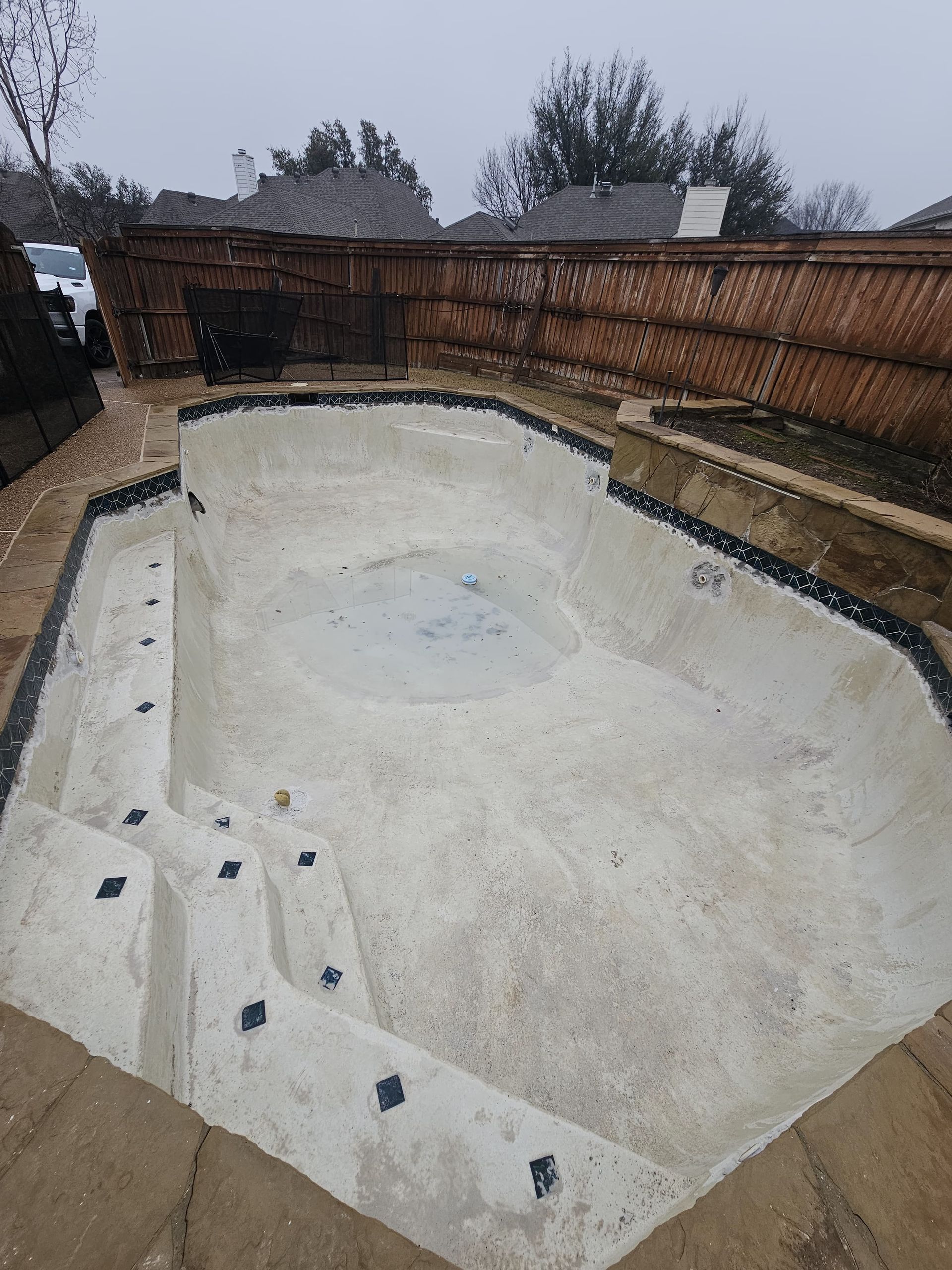 Empty swimming pool, light concrete, with stairs and dark tile accents, against a brick fence.
