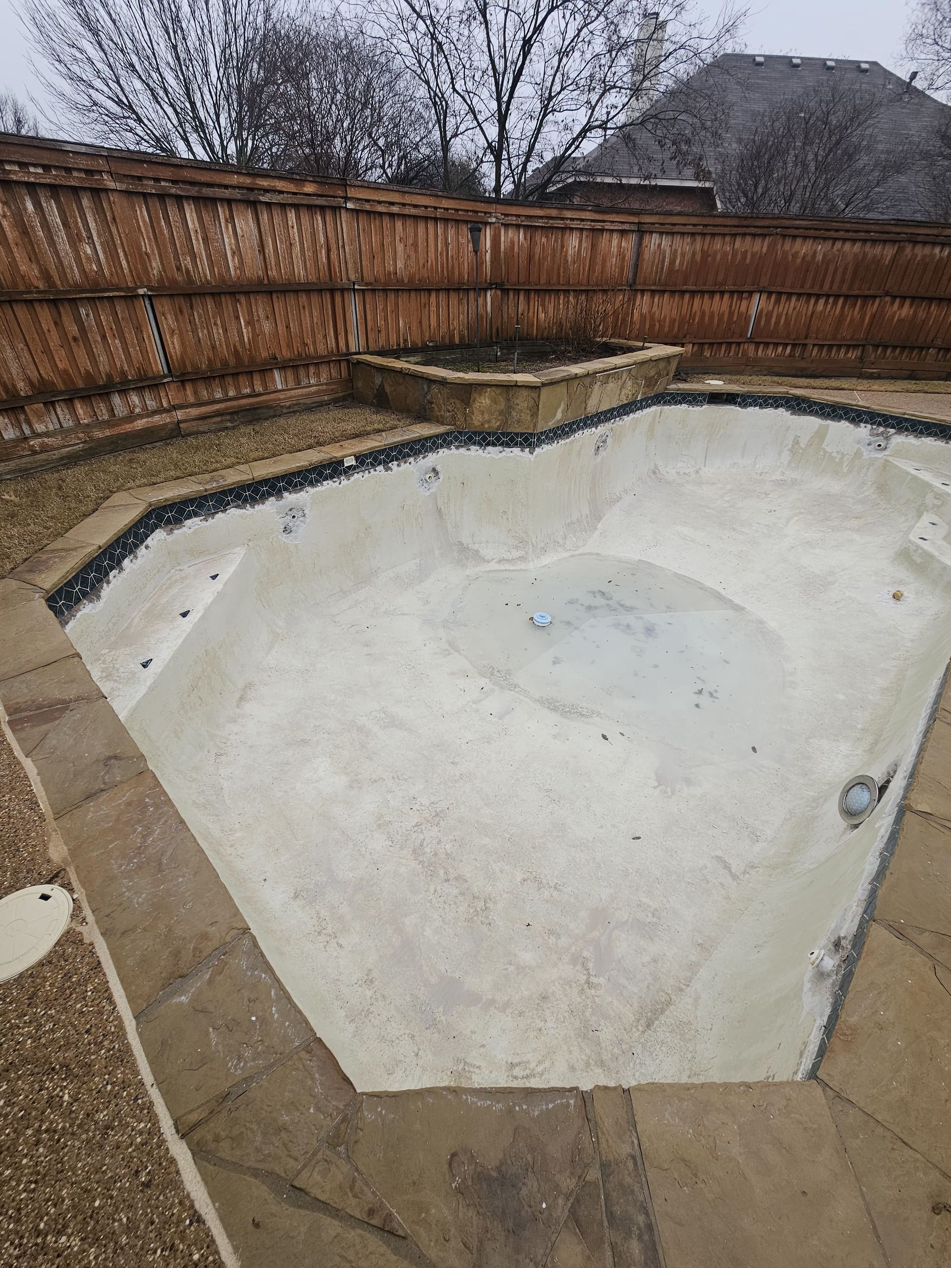 Empty, weathered swimming pool in a backyard, surrounded by stone and a wooden fence.
