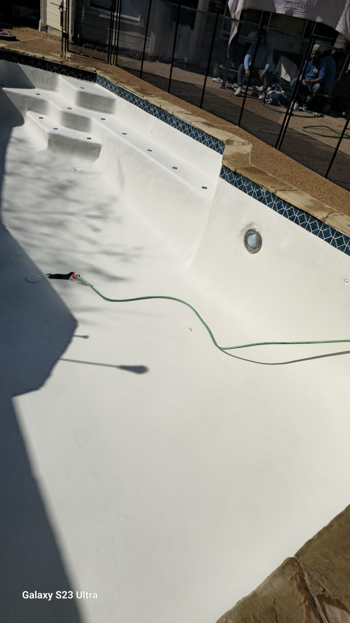 Newly painted white swimming pool with steps, surrounded by blue tile trim and a green hose.