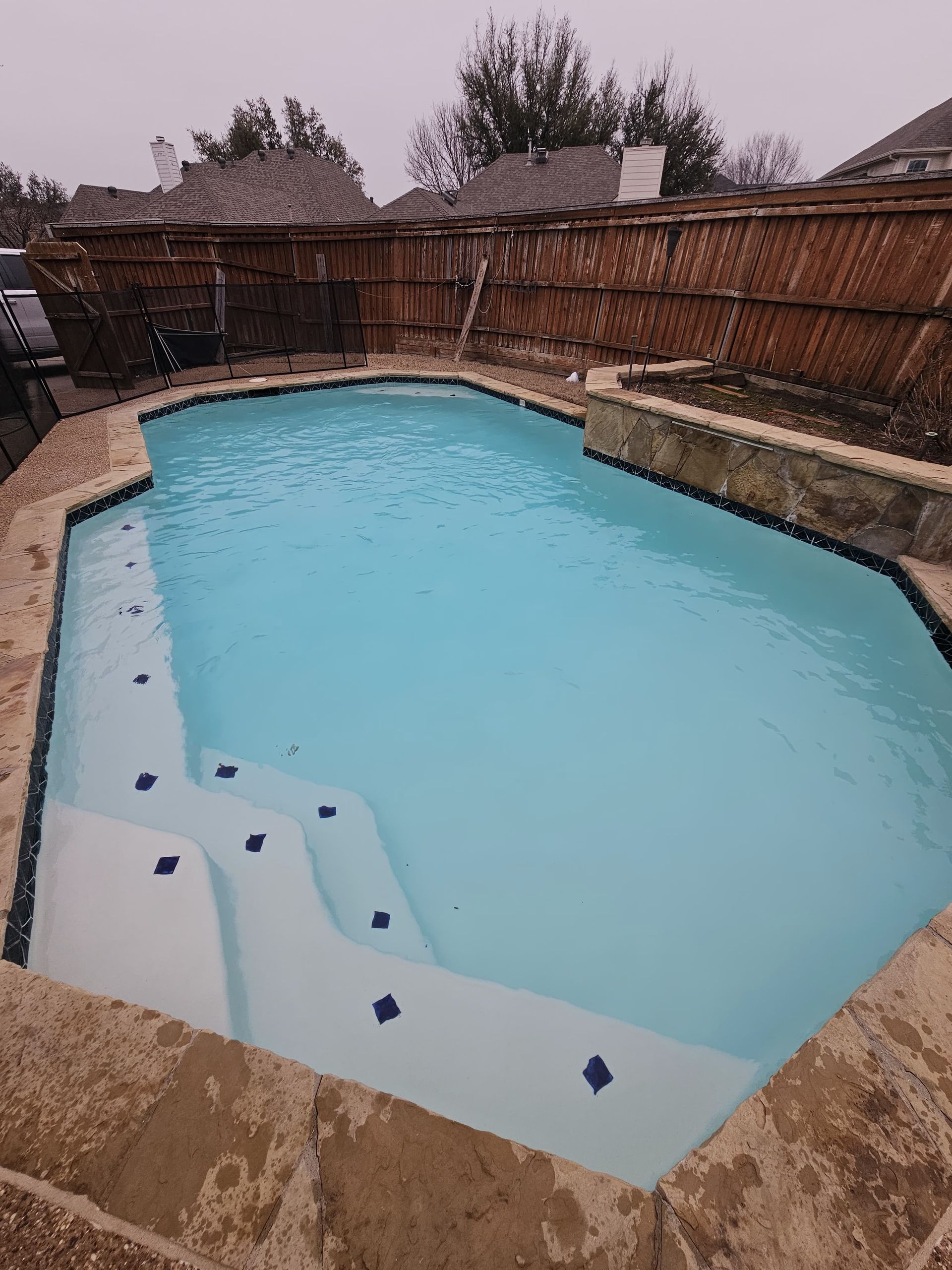 A swimming pool with clear blue water and steps, surrounded by stone and a wooden fence.