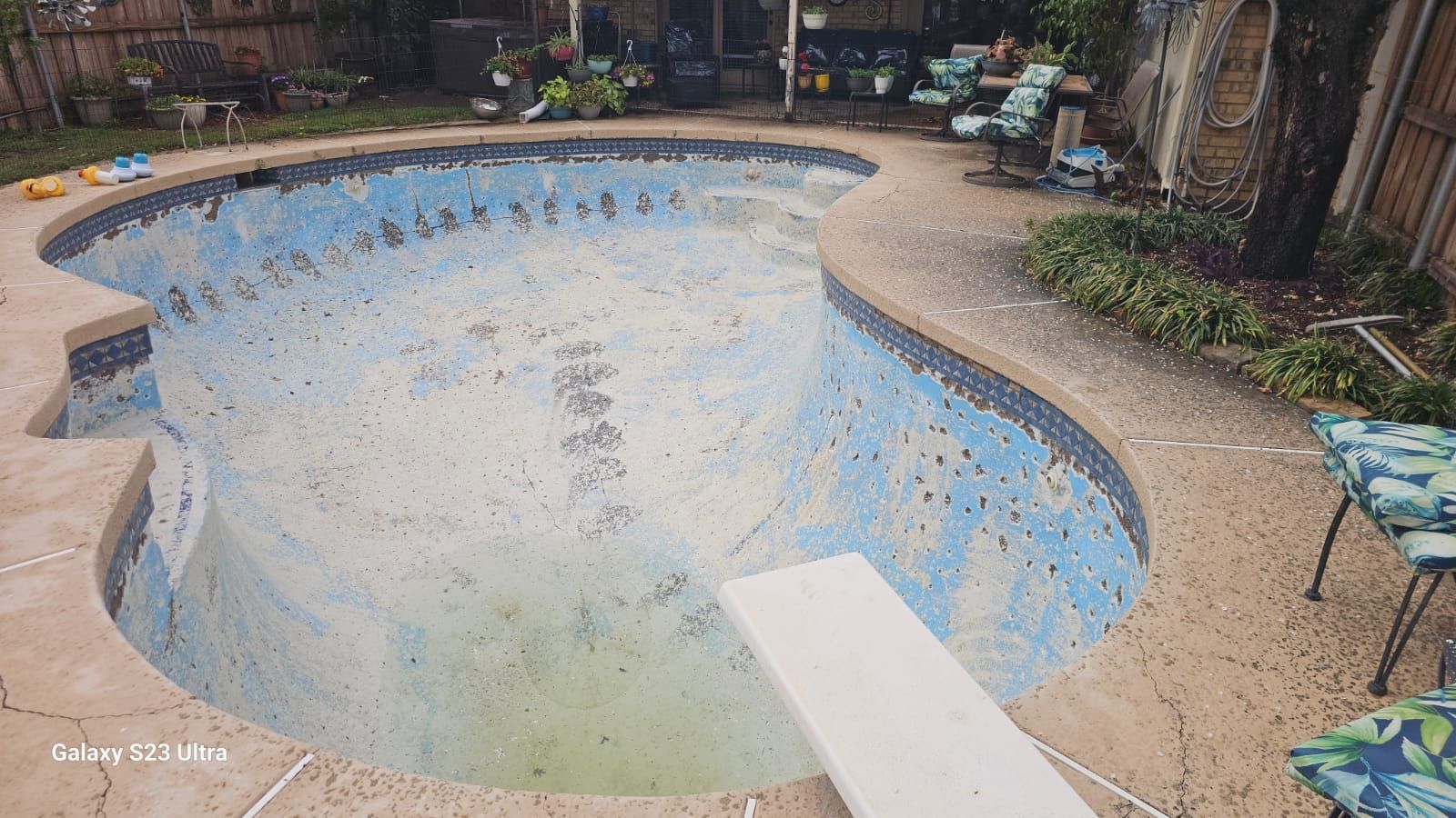 Empty swimming pool with peeling blue paint, diving board, and concrete deck. Backyard setting.