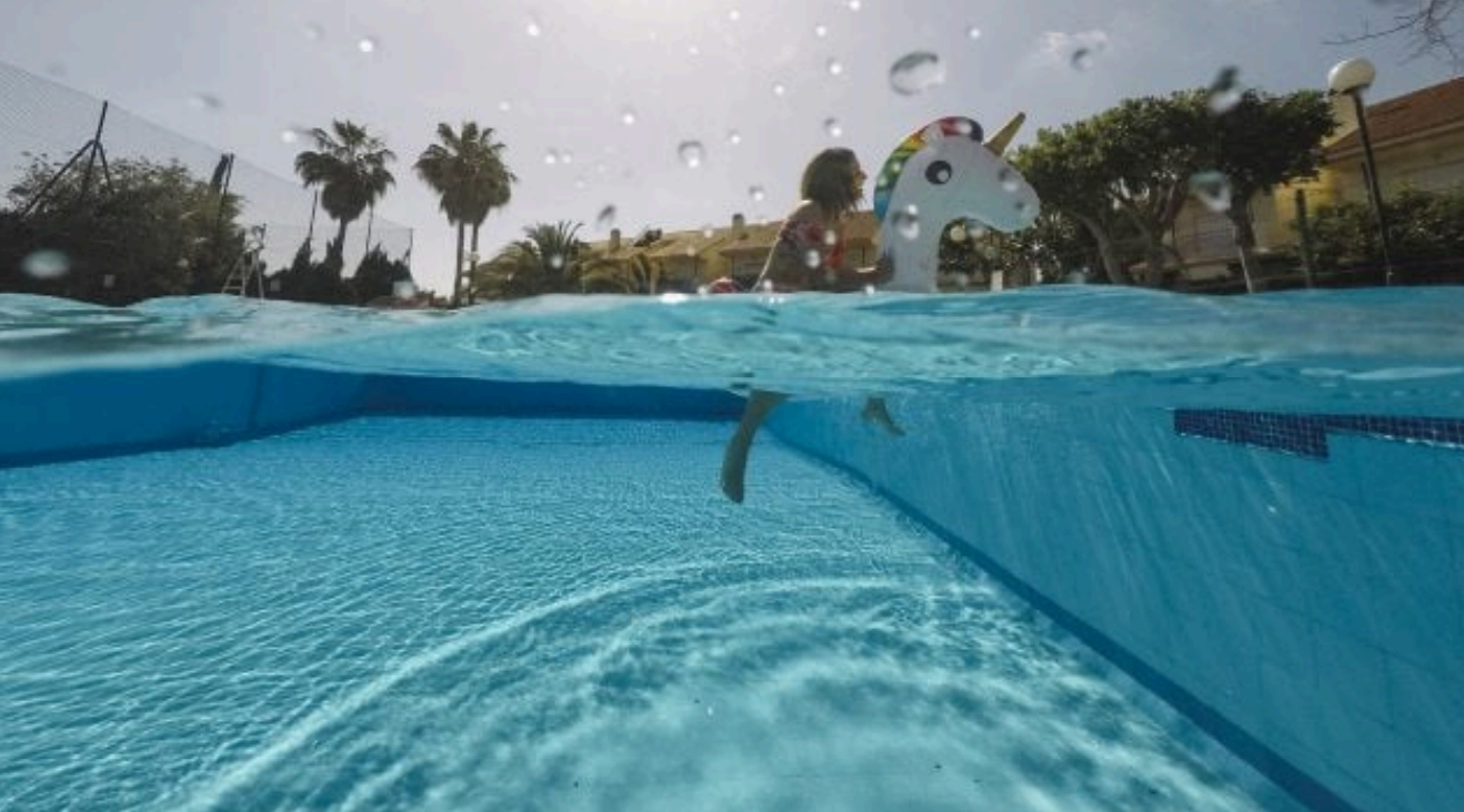 Split-level view of pool with child playing near a unicorn float. Palm trees and sunny sky in the background.