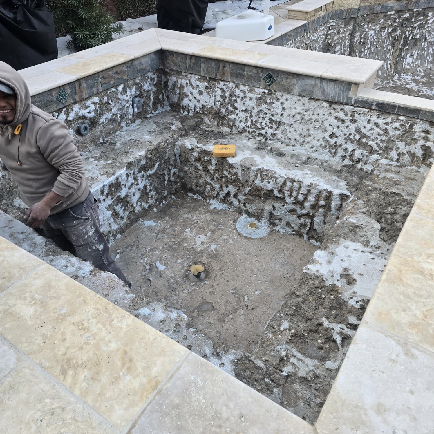 Man in a light brown hooded sweatshirt standing in a partially constructed spa. Travertine borders the spa's walls.