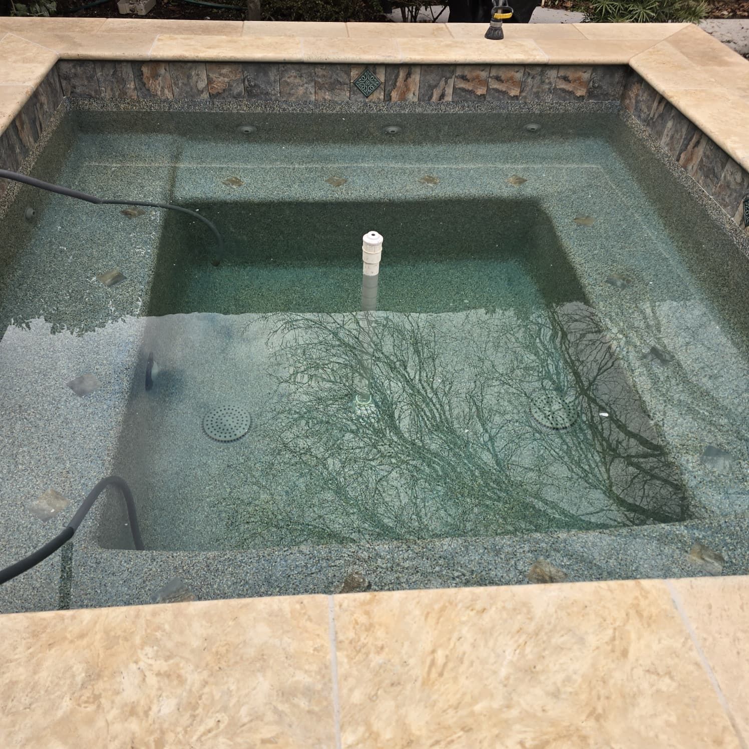 A rectangular spa with murky water, surrounded by light-colored tiles. A white pipe protrudes from the water.