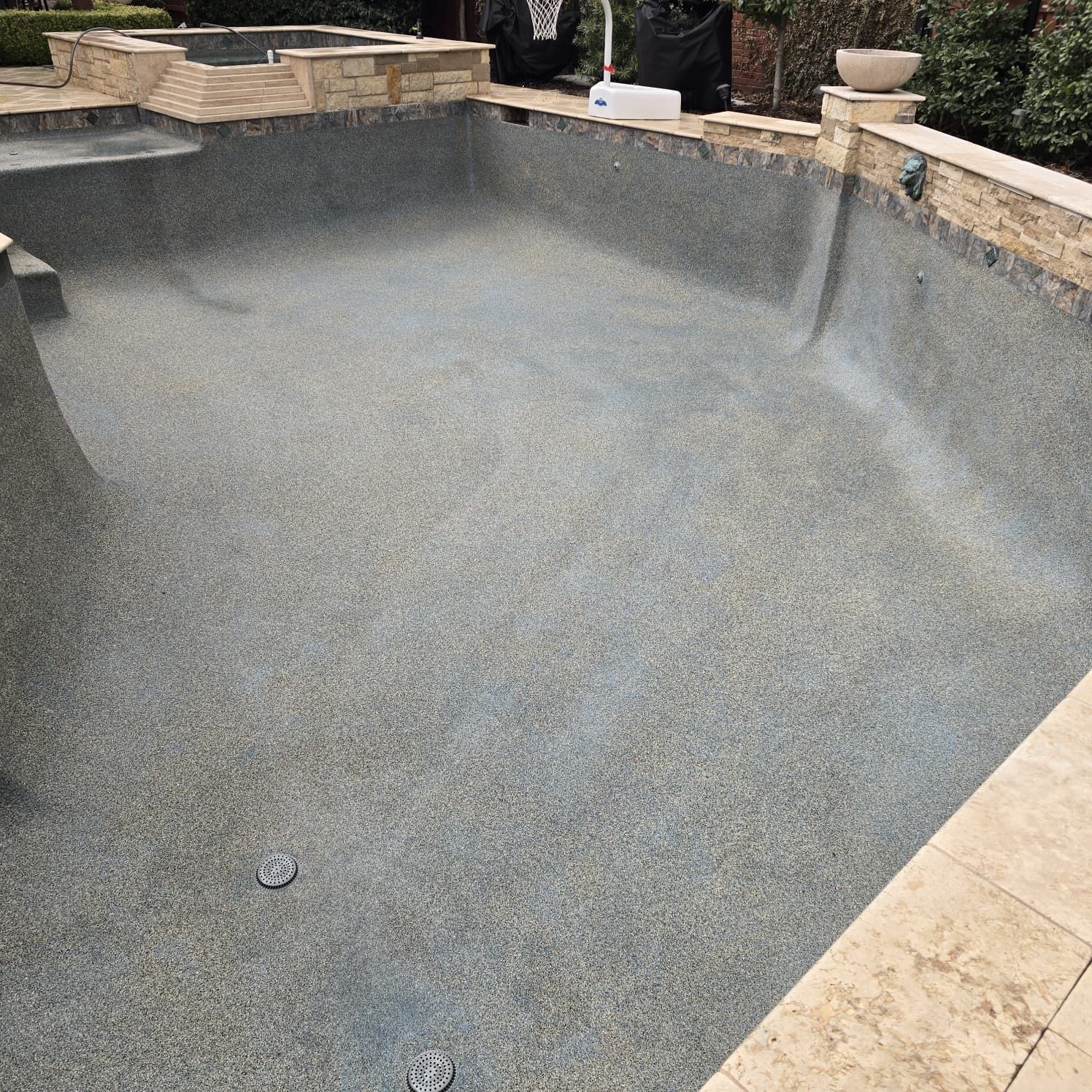 Empty swimming pool with speckled gray surface and tan stone coping.