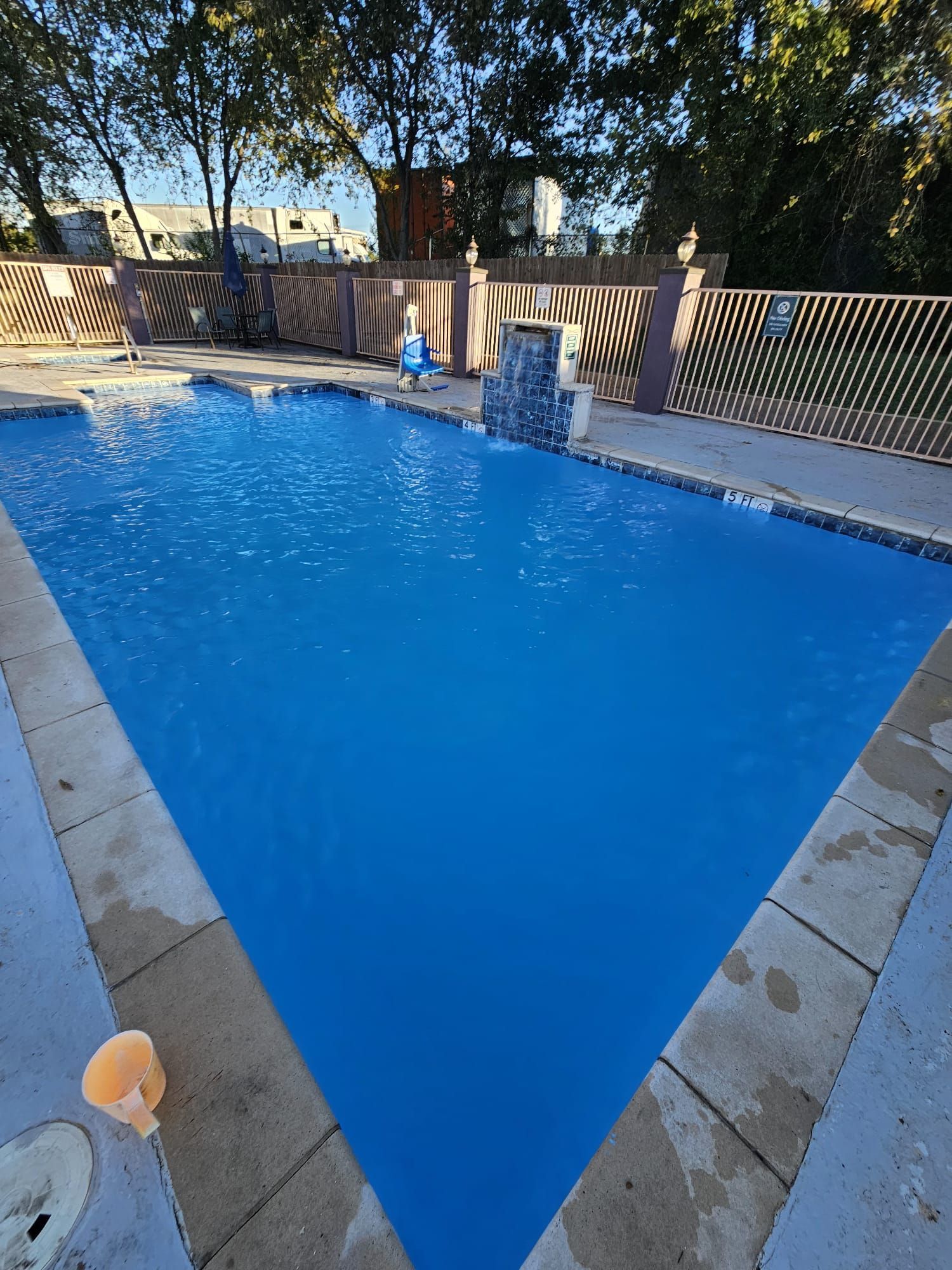 Blue swimming pool with waterfall, beige stone border, and a decorative fence.